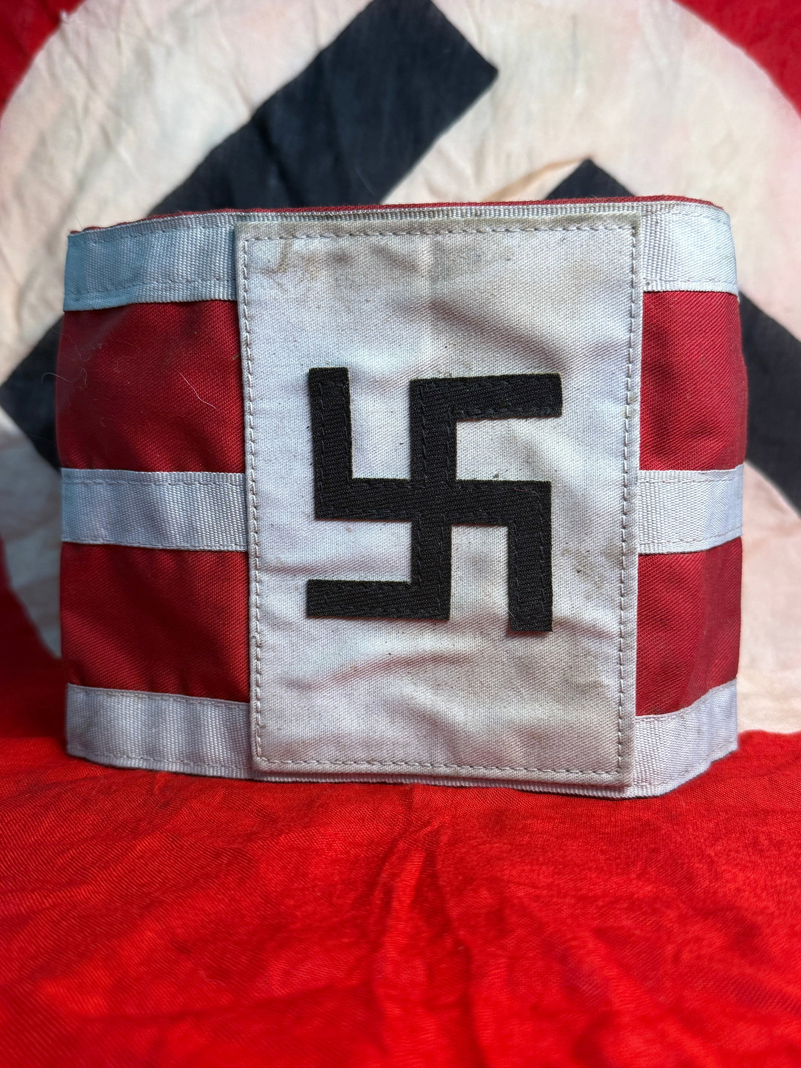 WWII German NSDAP Swastika Political Armband – Red, White & Black Cloth Construction: This WWII-era German political armband features the distinctive red field with contrasting white bands and a central white rectangular panel bearing a black embroidered swastika. The armband displays 