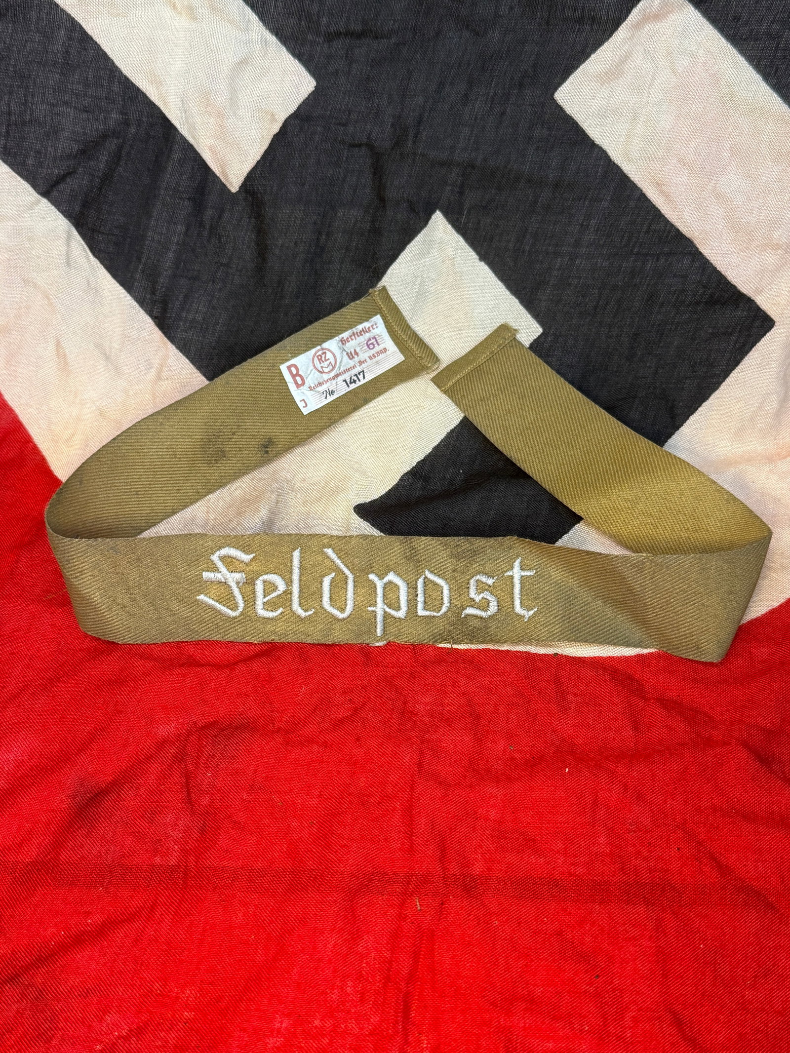 WWII German Feldpost Cuff Title with RZM Tag: This WWII German Feldpost cuff title is embroidered in silver thread on a field-gray woven band bearing the inscription "Feldpost." The reverse retains its original RZM paper control tag with printed 