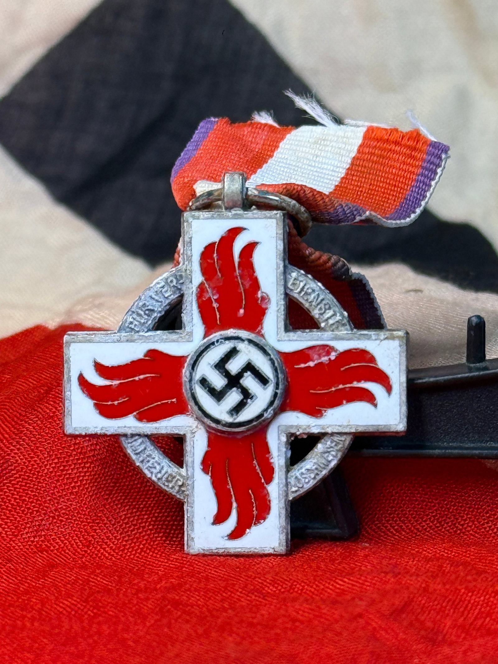 WWII German Fire Service Decoration 2nd Class Medal: This WWII German Fire Service Decoration 2nd Class Medal features a white enameled cross overlaid with red flame motifs and a central national emblem, suspended from its original ribbon. Instituted du