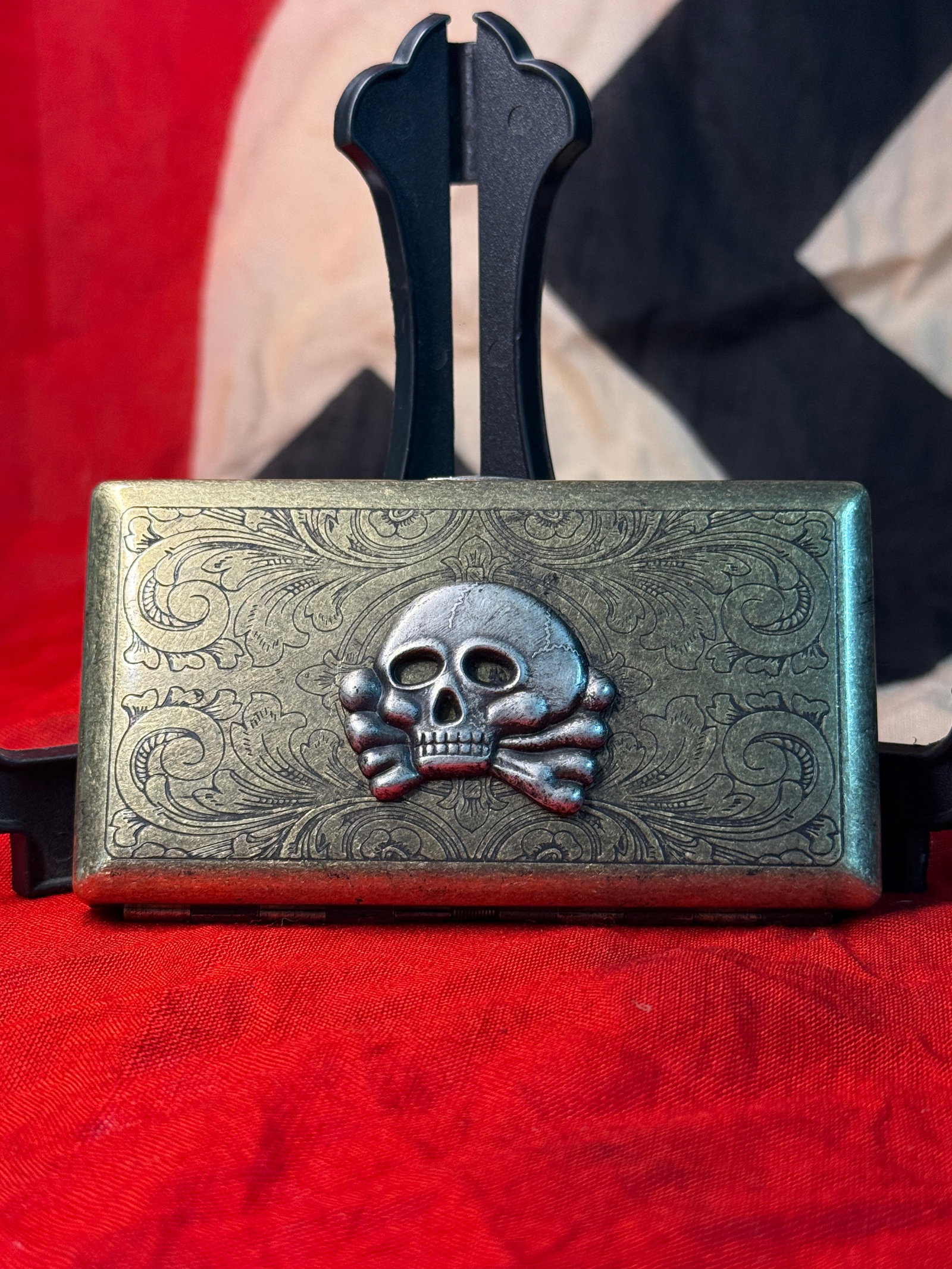 WWII German SS Totenkopf Cigarette Case: This WWII German cigarette case features an applied SS Totenkopf (Death's Head) skull and crossbones emblem mounted against an engraved foliate background on the front cover. The rectangular hinged me