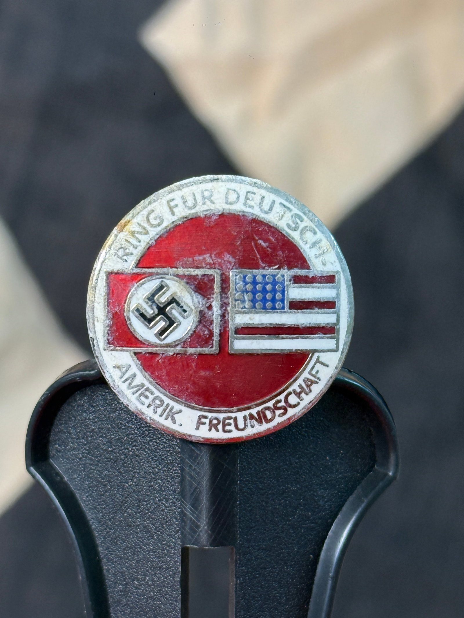 WWII German–American Friendship Lapel Badge By RZM M1/129: This WWII German–American Friendship lapel badge features crossed German and American flags against a red enamel background surrounded by the inscription "Ring für Deutsch-Amerik. Freundschaft" ("A
