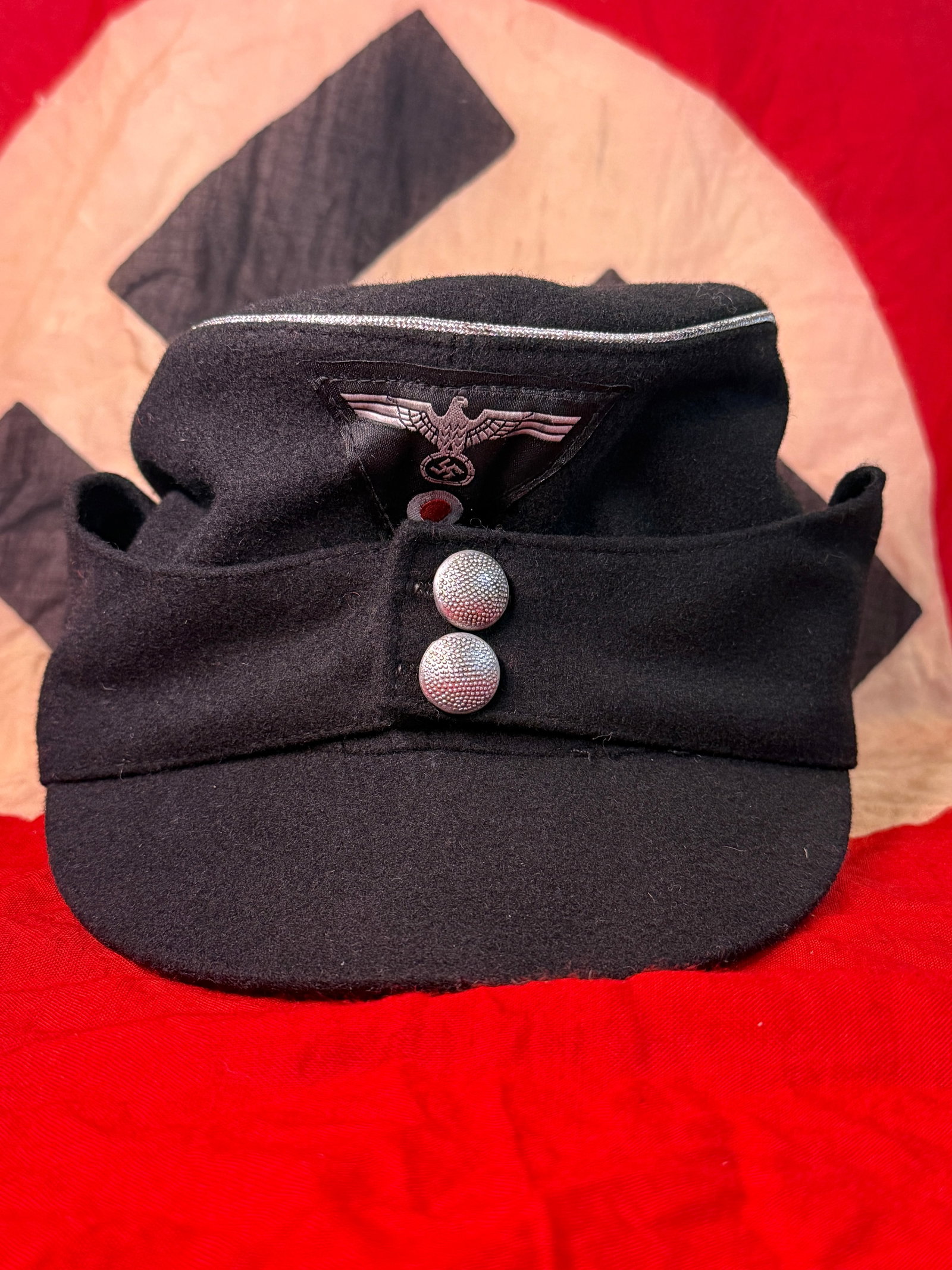 WWII German Waffen-SS M43 Field Cap: This WWII German Waffen-SS M43 field cap is constructed of black wool with silver piping, a machine-woven national eagle trapezoid insignia, and a pair of silver pebbled front buttons. The interior re