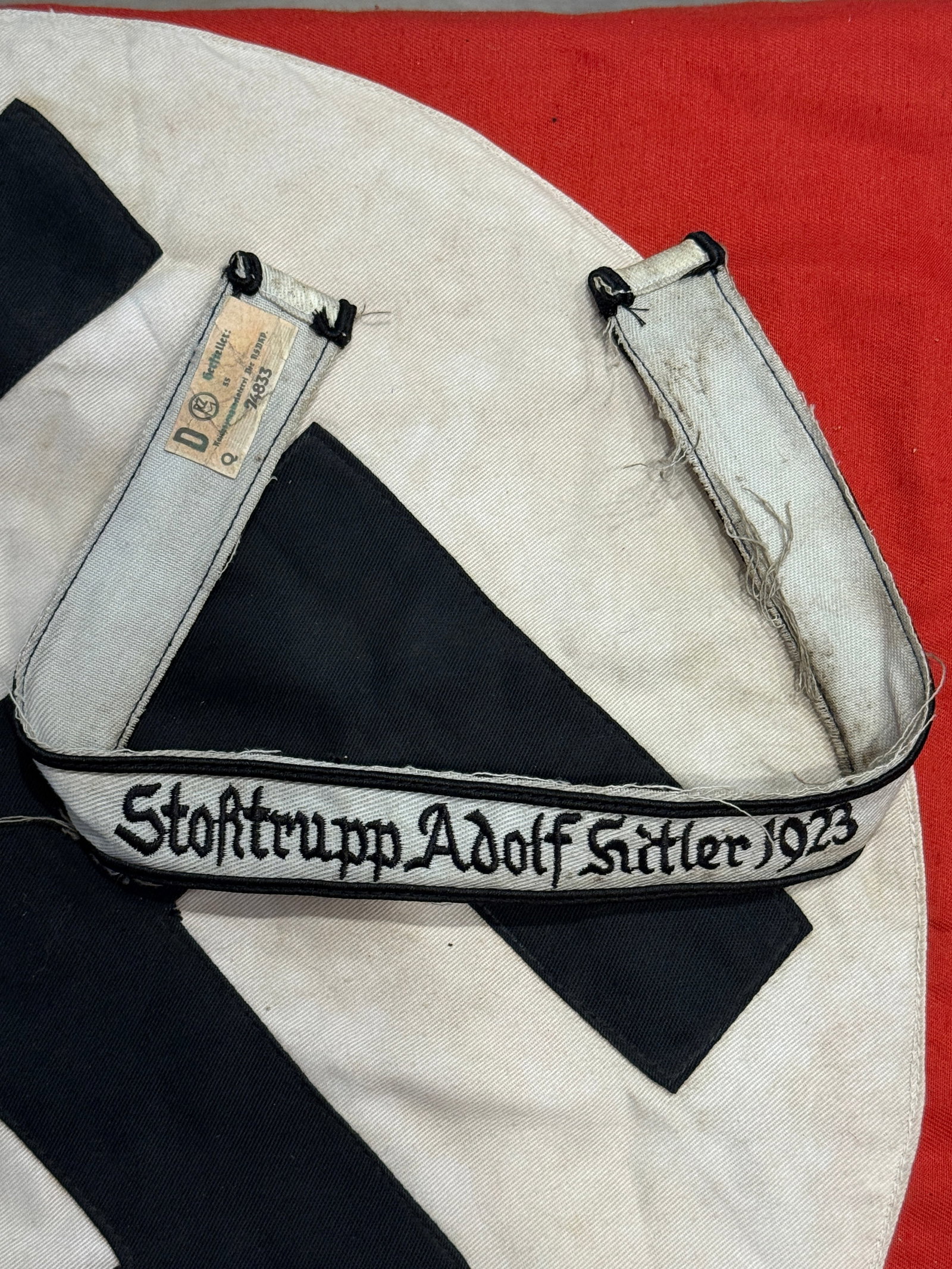 WWII German SA "Stosstrupp Adolf Hitler 1923" Cuff Title With RZM Paper Tag: This embroidered cuff title bears the inscription "Stosstrupp Adolf Hitler 1923" executed in black script on a silver-gray woven band with black edge piping. The reverse retains an RZM paper control l