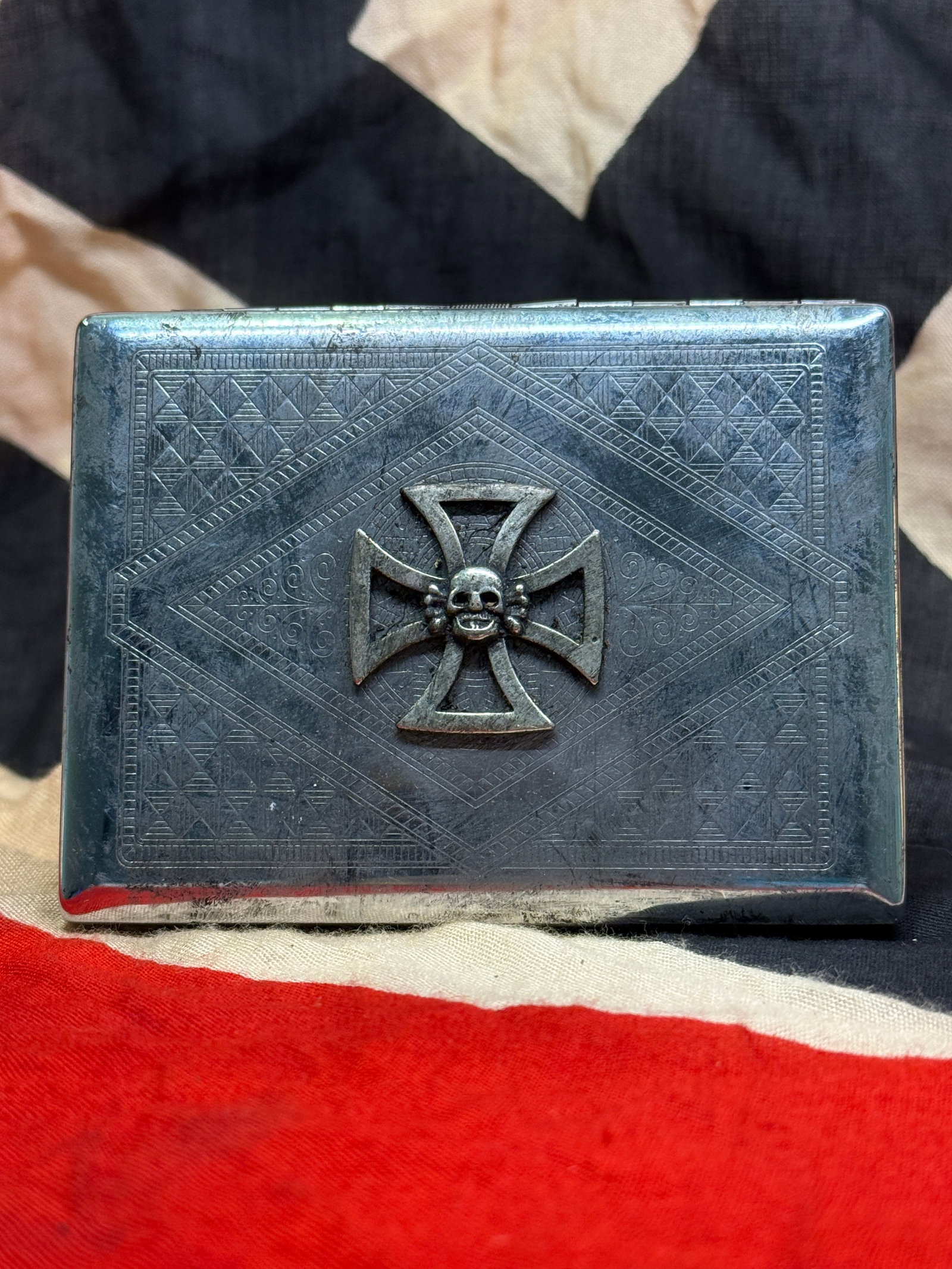 WWII German Totenkopf SS Officer Silver-Tone Cigarette Case – Skull & Iron Cross: This WWII German-style officer cigarette case features an elaborate silver-tone metal construction with finely engraved geometric and scrollwork decoration across the exterior. Mounted prominently to 