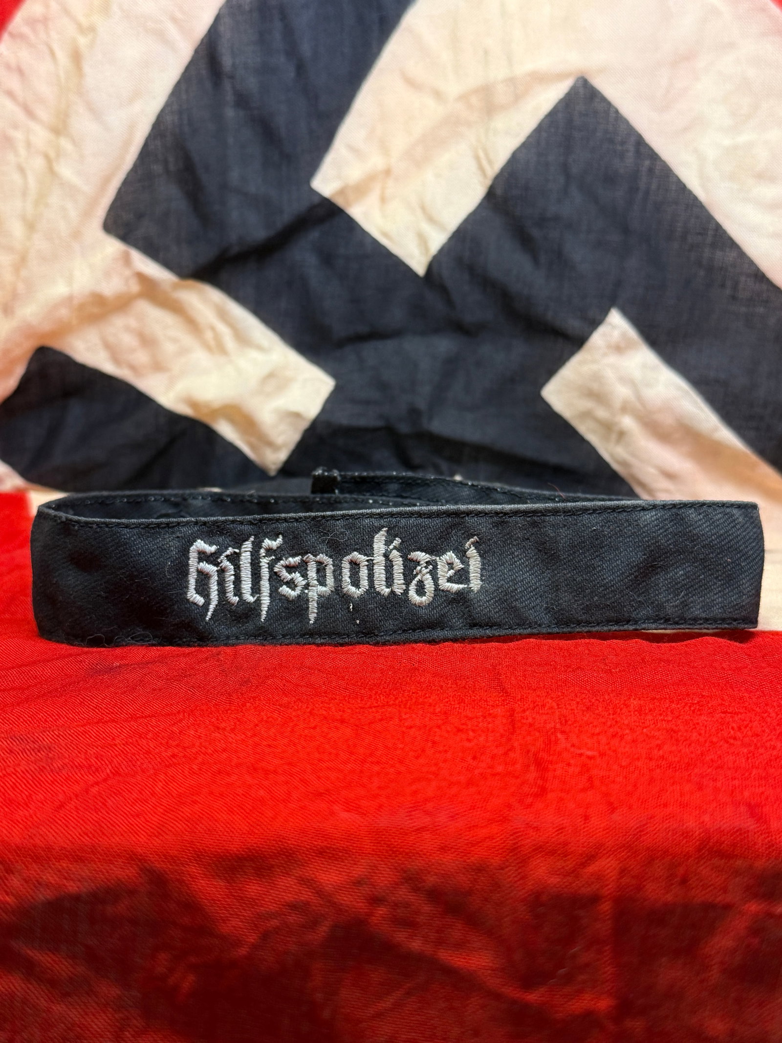 WWII German "Hilfspolizei" Armband: This black cloth armband is embroidered in silver-gray thread with the word "Hilfspolizei," identifying the wearer as a member of the German Auxiliary Police. The reverse displays period stitching and