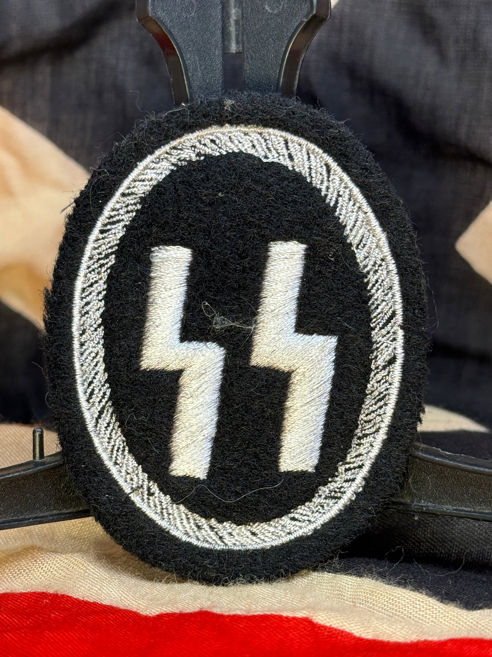 WWII German Waffen-SS Sleeve Patch With RZM Paper Tag: This oval black cloth sleeve patch features the embroidered white Waffen-SS double Sig runes enclosed within a silver twisted border. The reverse retains an RZM paper control label bearing approval ma