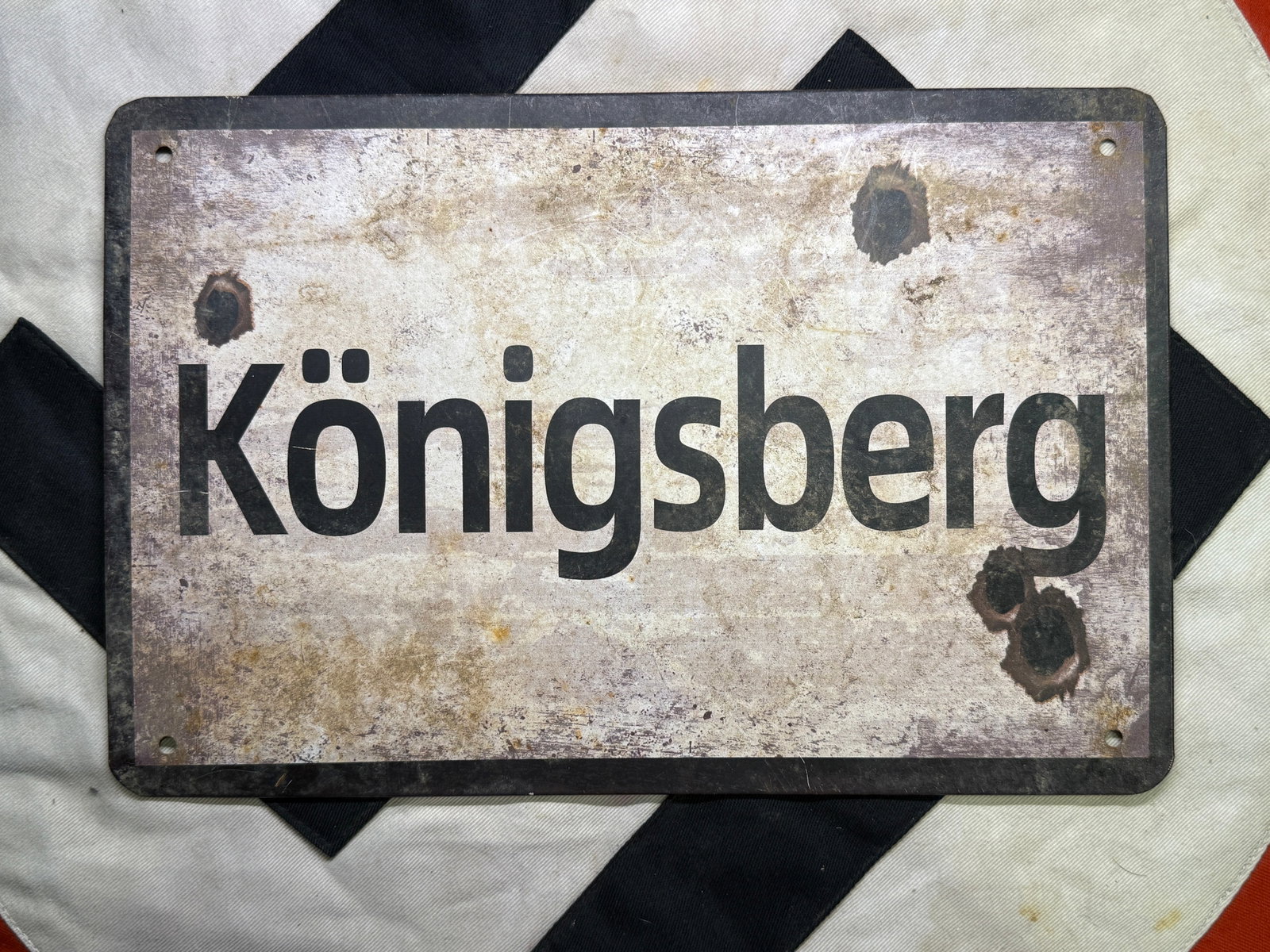 WW2 German Königsberg Place-Name Display Plaque: Rectangular pressed-sheet-metal plaque bearing the German place name “Königsberg” in bold black lettering. The face has a pale, intentionally weathered finish with dark simulated impact marks