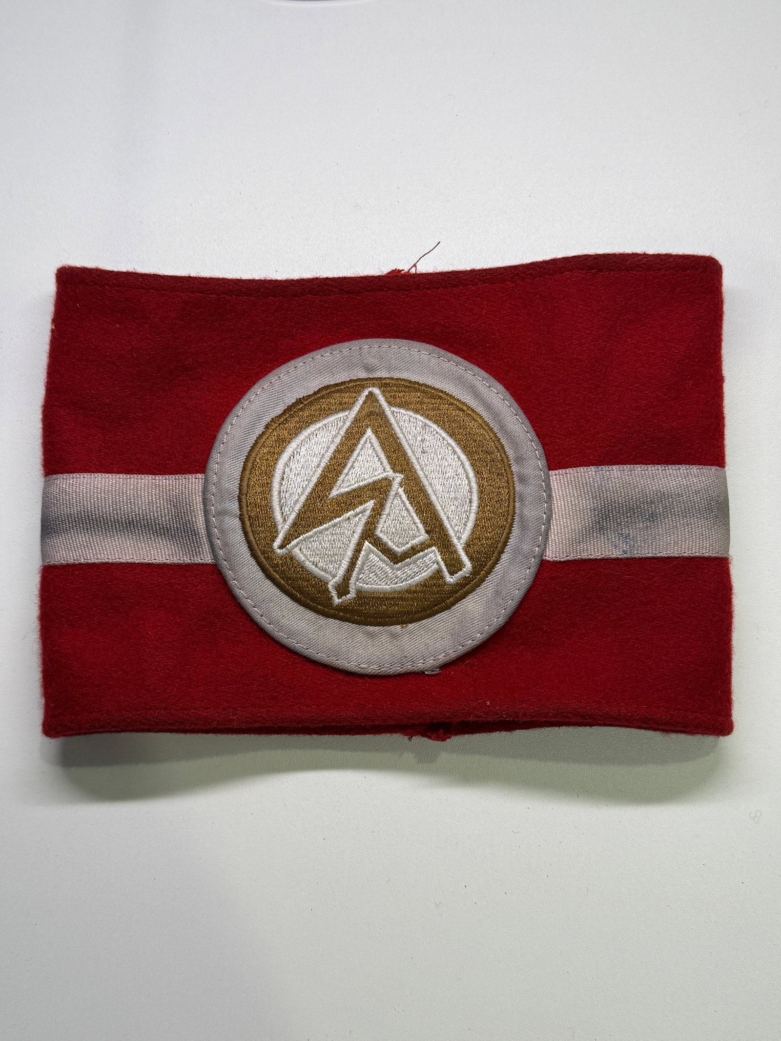 WWII German SA Wehrmannschaft Sports Armband with RZM Label: Red wool armband associated with the Sturmabteilung’s military-training and athletic program. A horizontal silver-gray woven band crosses the center, supporting a circular applied insignia worked in