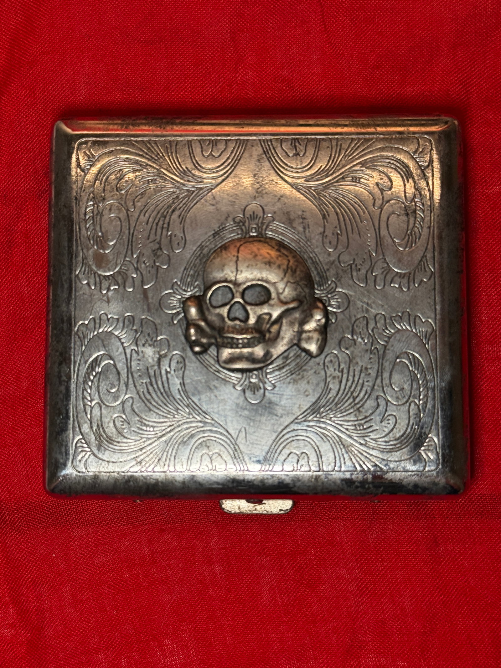 WWII German SS Totenkopf Skull Silver-Tone Engraved Cigarette Case: This WWII German SS-style cigarette case features an elaborate engraved silver-tone metal body with a prominent raised Totenkopf skull and crossbones emblem mounted at the center of the front. The ext