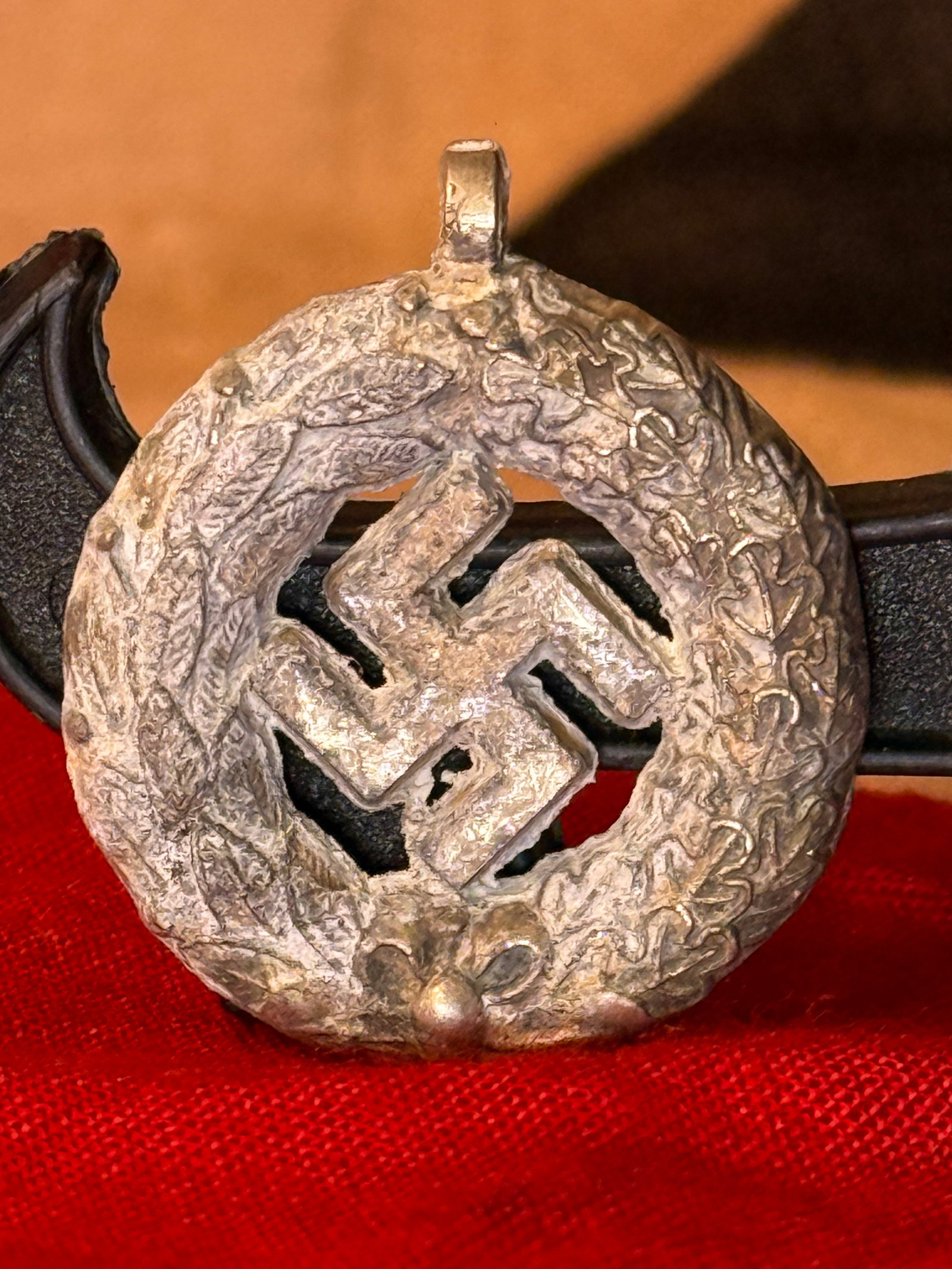 WWII German Swastika and Oak-Leaf Wreath Pendant: Circular silver-colored German political pendant formed with an openwork central swastika surrounded by a raised wreath of oak leaves and acorns. A rectangular suspension loop projects from the upper 