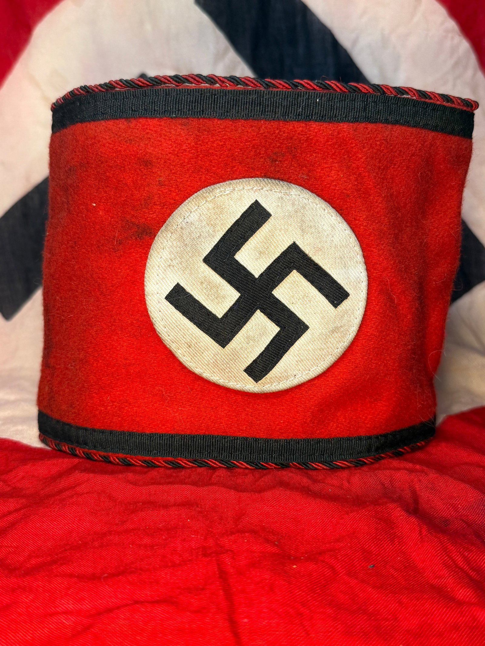 WWII German Waffen SS Wool Armband with RZM Label: Red wool armband centered with a sewn white circular field bearing a black swastika. Black woven bands run along the upper and lower borders, accompanied by twisted red-and-black piping that gives the