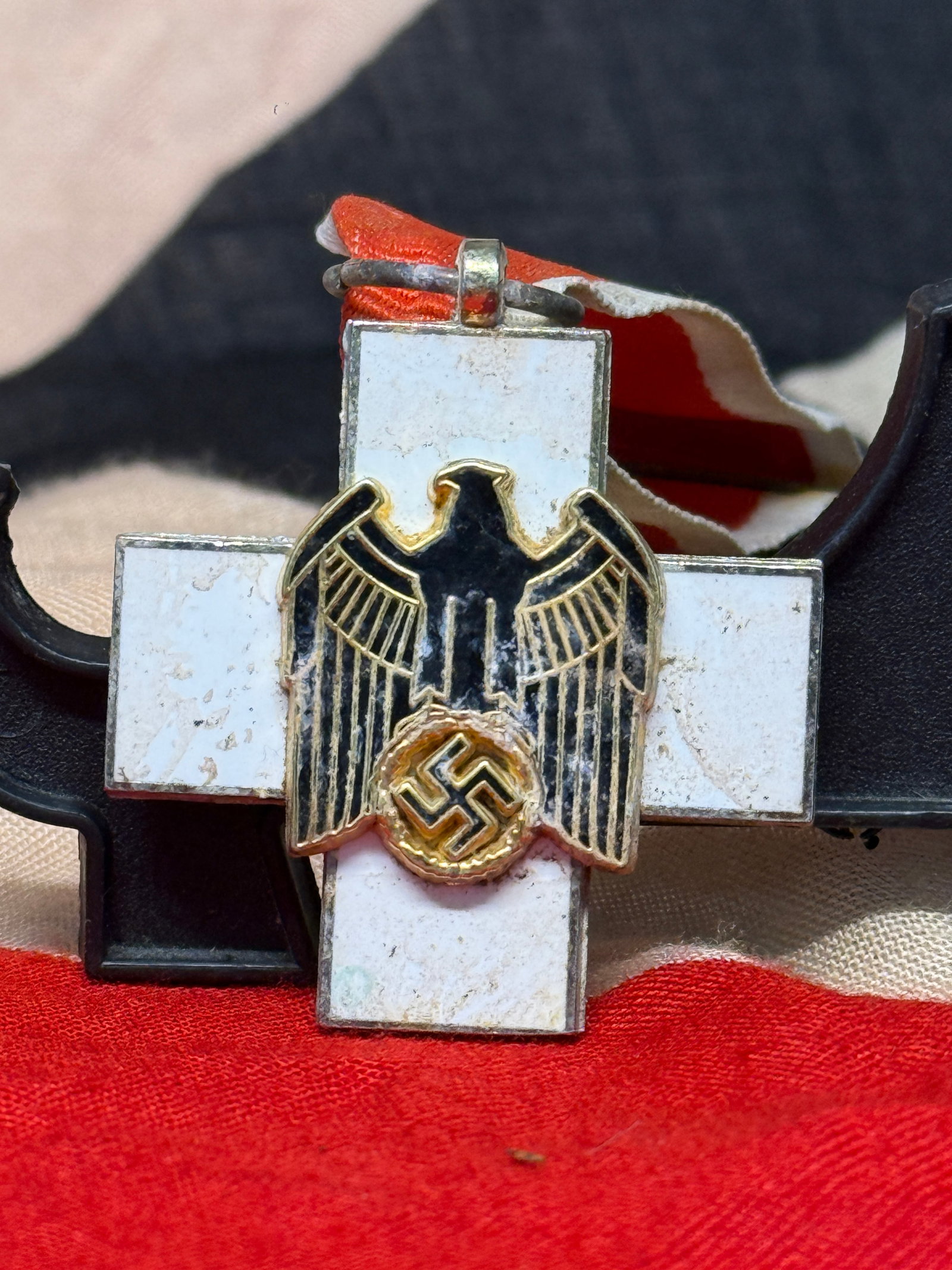 WWII German Social Welfare Decoration, Third Class: White enamel cross-form decoration with a black enamel national eagle clutching a wreath-and-swastika device at its center, suspended from a red-and-white ribbon. This Third Class Social Welfare Decor