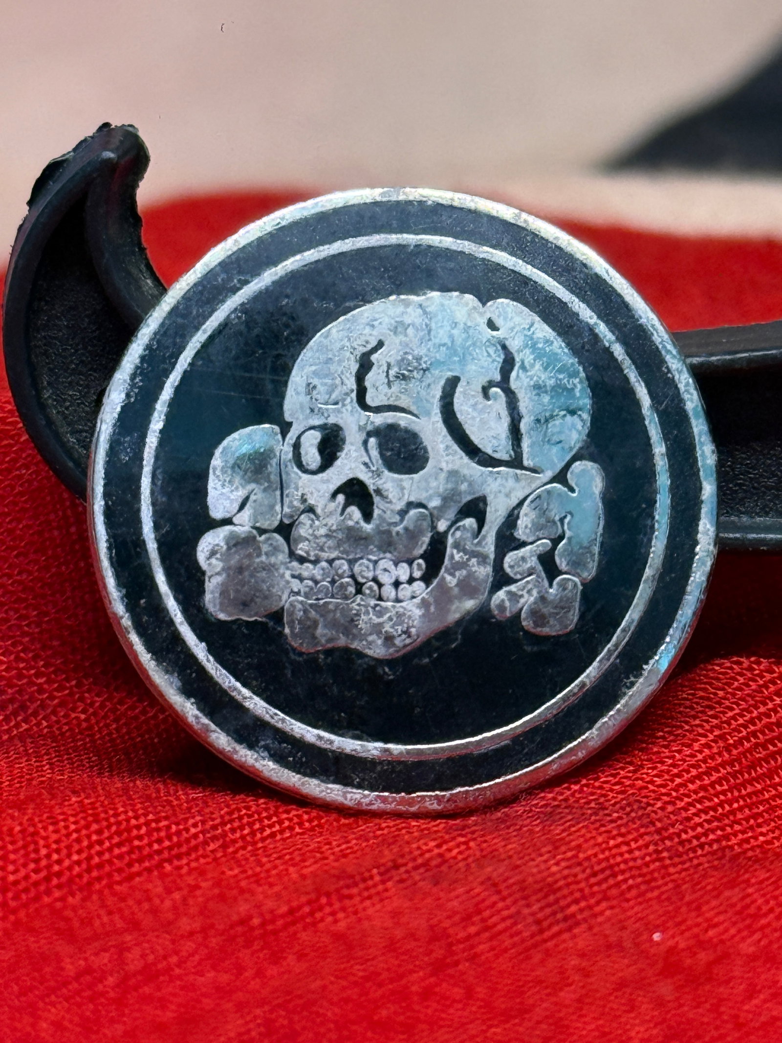 WWII German SS Totenkopf Skull-and-Crossbones Badge: Circular metal badge displaying a raised skull and crossed bones against a dark field, enclosed by a narrow outer border. The reverse retains its pin fitting. The Totenkopf motif was closely associate