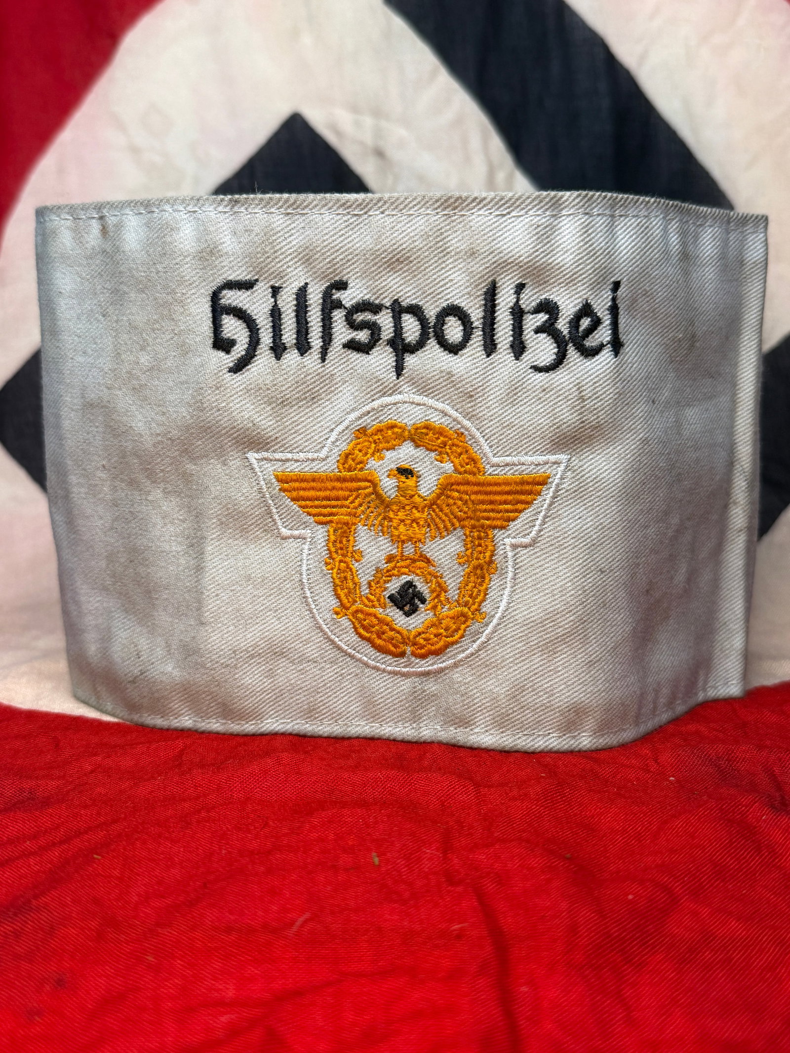 WWII German Nazi-Era Hilfspolizei Auxiliary Police Armband with RZM Tag: White cloth armband embroidered in black with “Hilfspolizei,” or Auxiliary Police, above a gold-toned police-style eagle and wreath insignia with a central swastika. The armband is constructed