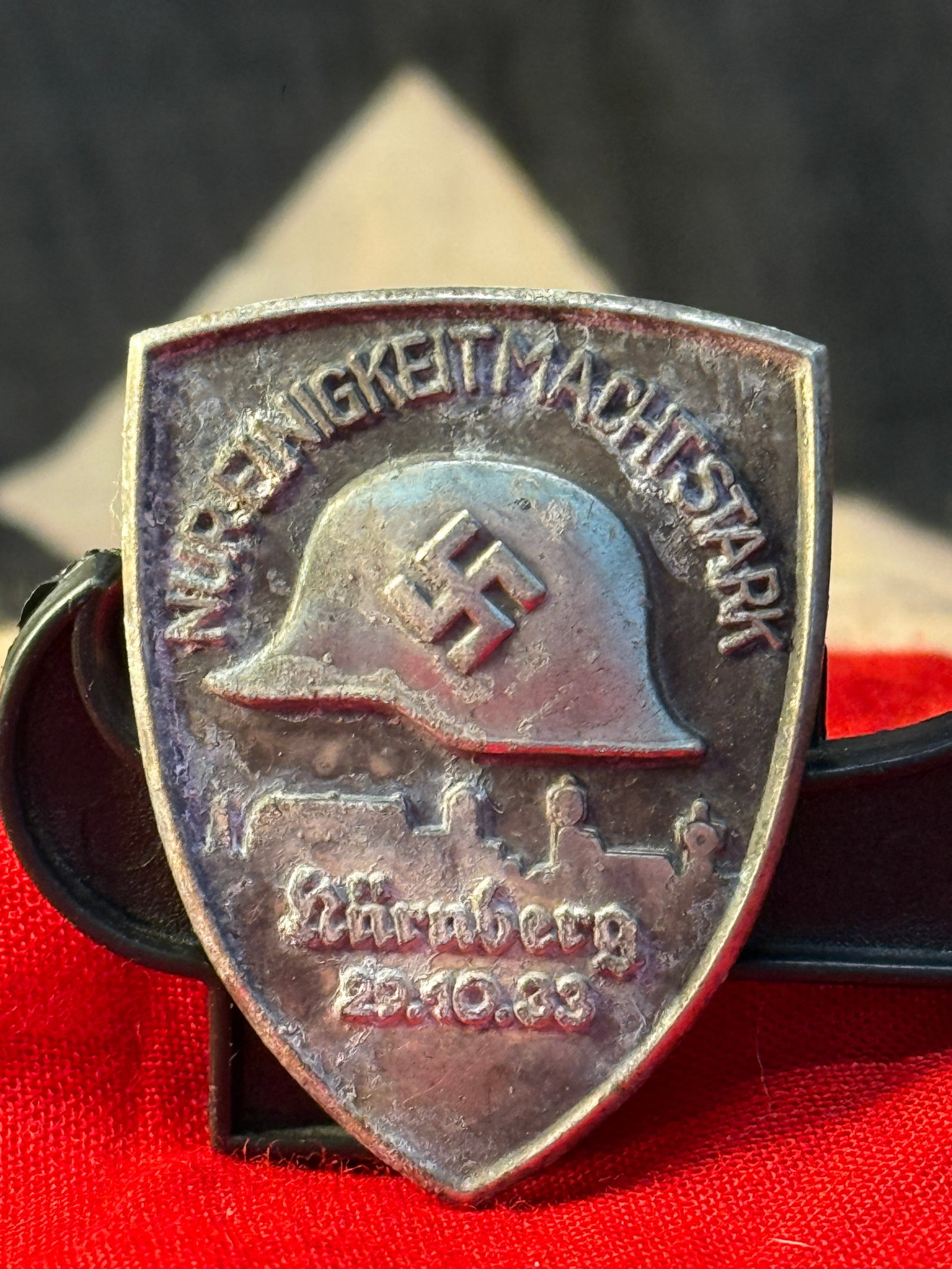 WWII German Winter Relief (WHW) Nuremberg Tinnie – Reichsparteitag Nürnberg 23.10.33: This die-struck commemorative tinnie bears the inscription "Wir Tragen Mit Stolz Hakenkreuz" above a steel helmet and the wording "Nürnberg 23.10.33," commemorating an early National Socialist gather