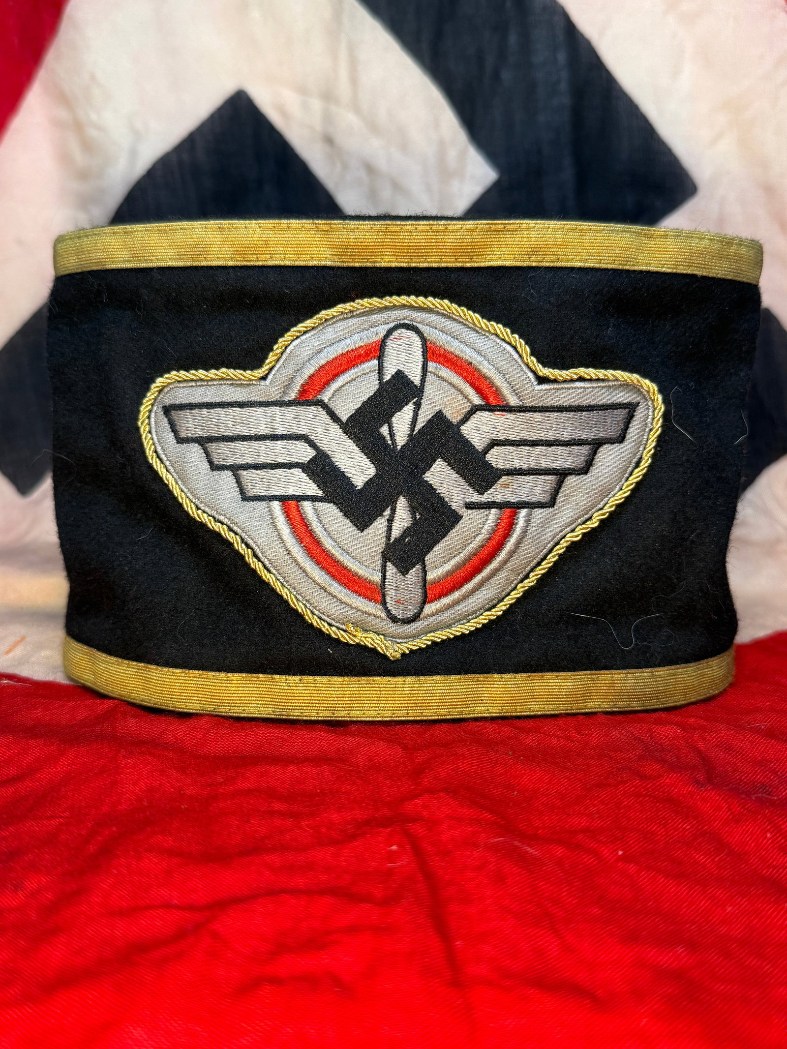 WWII German NSKK Leader's Armband: This black wool armband features the embroidered NSKK emblem consisting of a winged steering wheel with a vertical sword passing through the center, enclosed within a red ring and finished with gold-c
