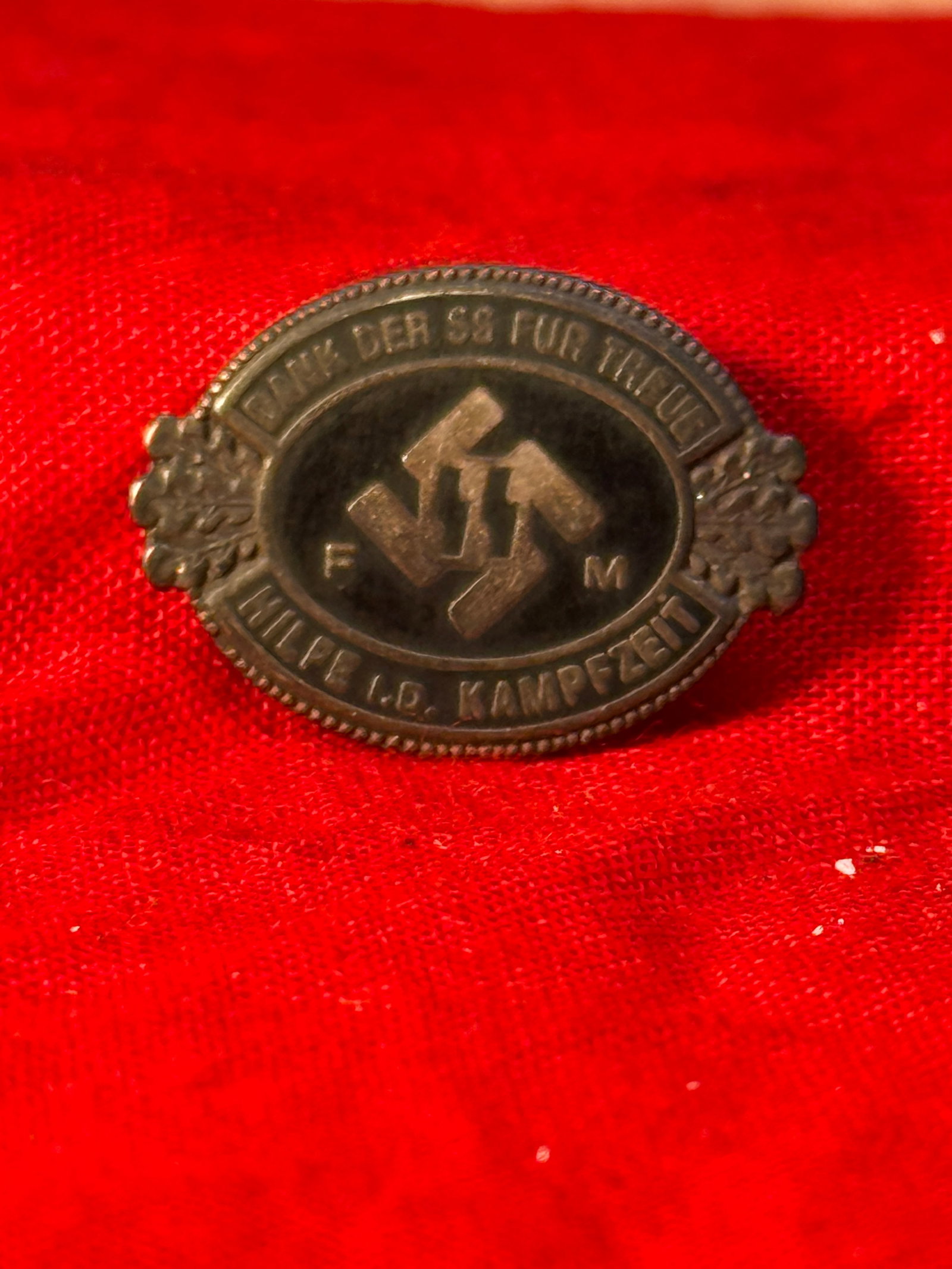 WWII German SS-FM Honor Pin By RZM M1/133: This oval metal honor pin features the intertwined FM monogram incorporating a swastika, surrounded by the German inscription "Kämpft der SS für Tallinn" with decorative oak leaf accents at each sid