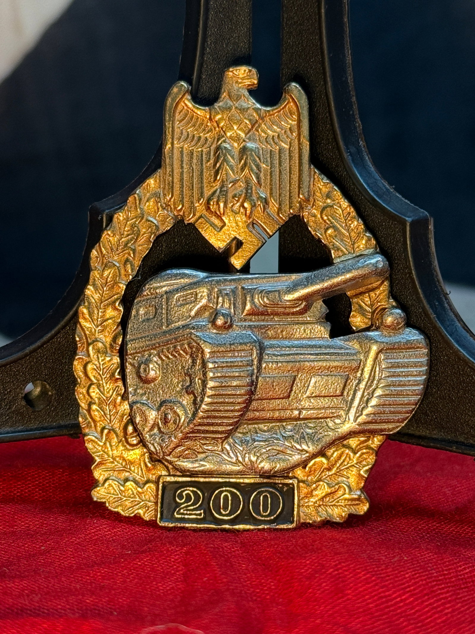 WWII German Panzer Assault Badge with "200" Tank Engagement Device: This gold-finished badge depicts a Panzer tank beneath the national eagle and oak leaf wreath, with a black enamel "200" engagement device mounted along the base. The reverse retains its vertical pinb
