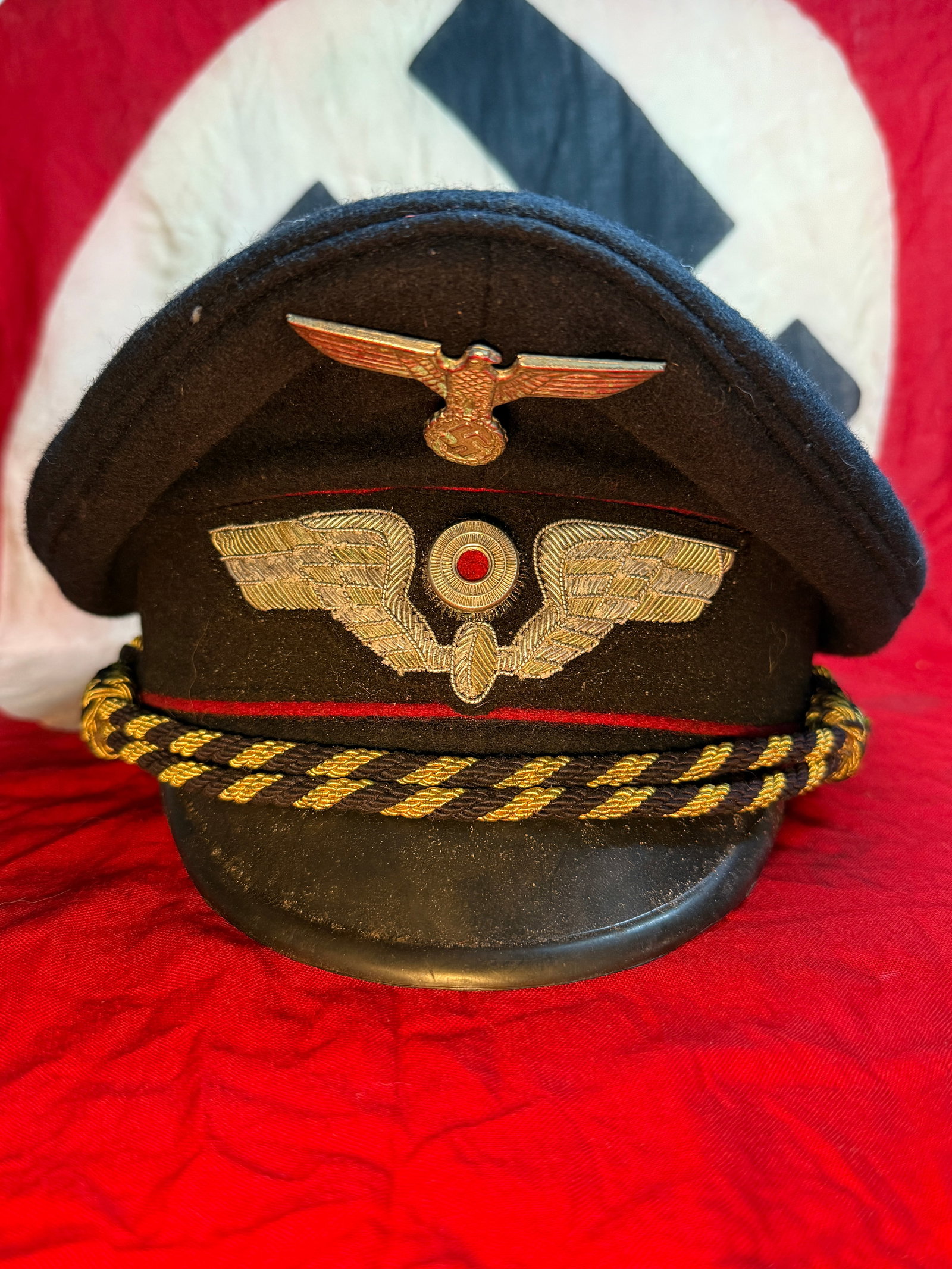 WWII German NSDAP Political Leader Visor Cap: This black wool visor cap is fitted with a metal national eagle, finely embroidered political wreath cockade with red center, black-and-gold bullion chin cord, and red piping characteristic of NSDAP P