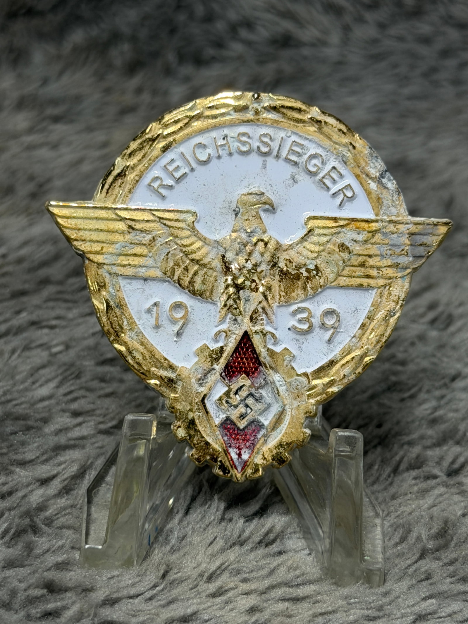 WWII German 1939 Reichssieger Hitler Youth National Competition Award Badge – G. Brehmer: A striking WWII German Reichssieger award badge dated 1939, featuring a large gilt eagle with outstretched wings positioned across a white enameled circular field bearing the inscription “REICHSSIEG