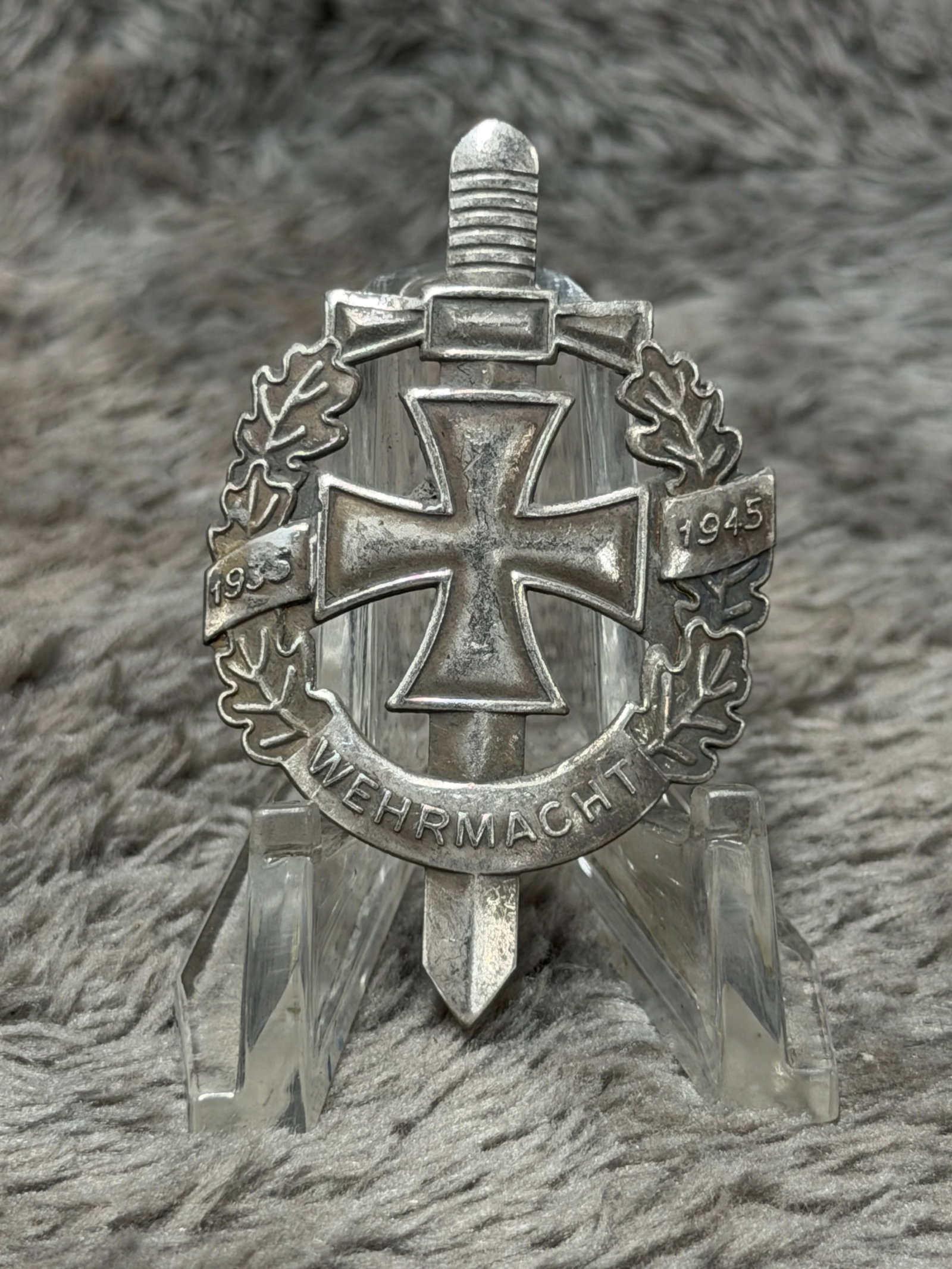 WWII German Wehrmacht 1935–1945 Iron Cross & Sword Veteran Commemorative Badge: A visually impressive WWII German Wehrmacht commemorative badge constructed around a vertical sword and prominent Iron Cross, surrounded by a detailed oak-leaf wreath. Small banners positioned across 