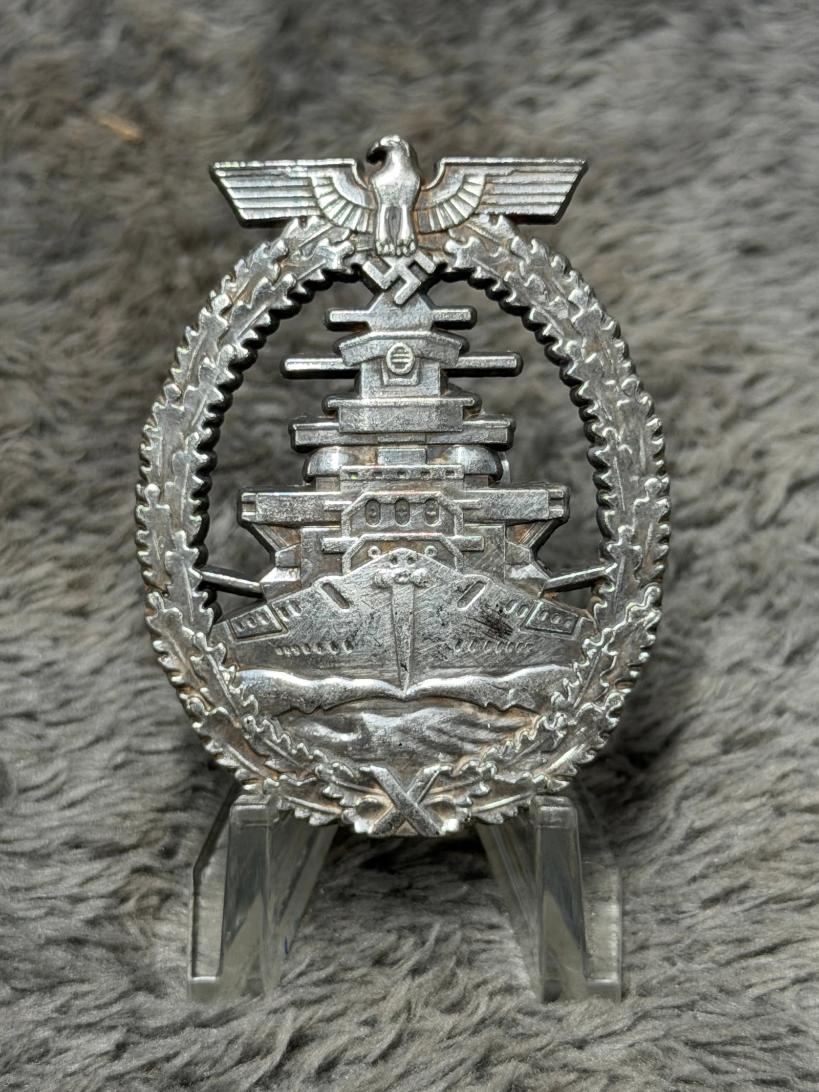 WWII German Kriegsmarine High Seas Fleet War Badge – Fec. Adolf Bock, Schwerin Berlin: A visually impressive WWII German Kriegsmarine High Seas Fleet War Badge featuring a detailed frontal depiction of a German battleship cutting through stylized ocean waves, surrounded by an oval oak-l