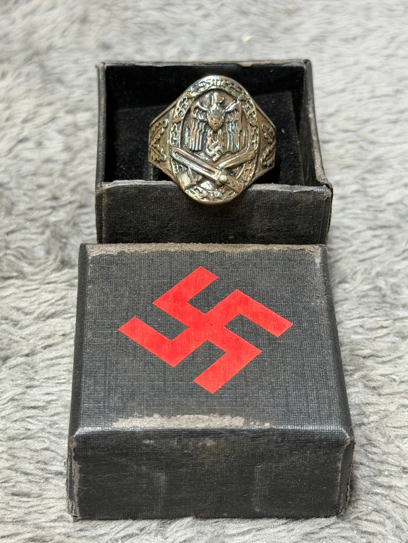 WWII German Army Heer Infantry Assault Combat Ring with Eagle, Crossed Weapons & Oak Leaves: WWII German military-themed finger ring featuring an elaborate raised combat motif across its large shield-shaped face. The central design depicts a German eagle above a prominent swastika, framed by 