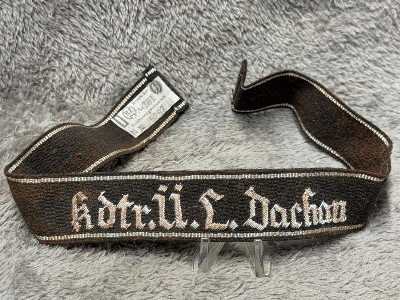 WWII German SS Dachau Concentration Camp “Kdr. Ü. L. Dachau?? Cuff Title with RZM Label: WWII German SS cuff title associated with the Dachau concentration camp, constructed from dark woven fabric with silver-colored Gothic embroidered lettering reading “Kdr. Ü. L. Dachau” and bo