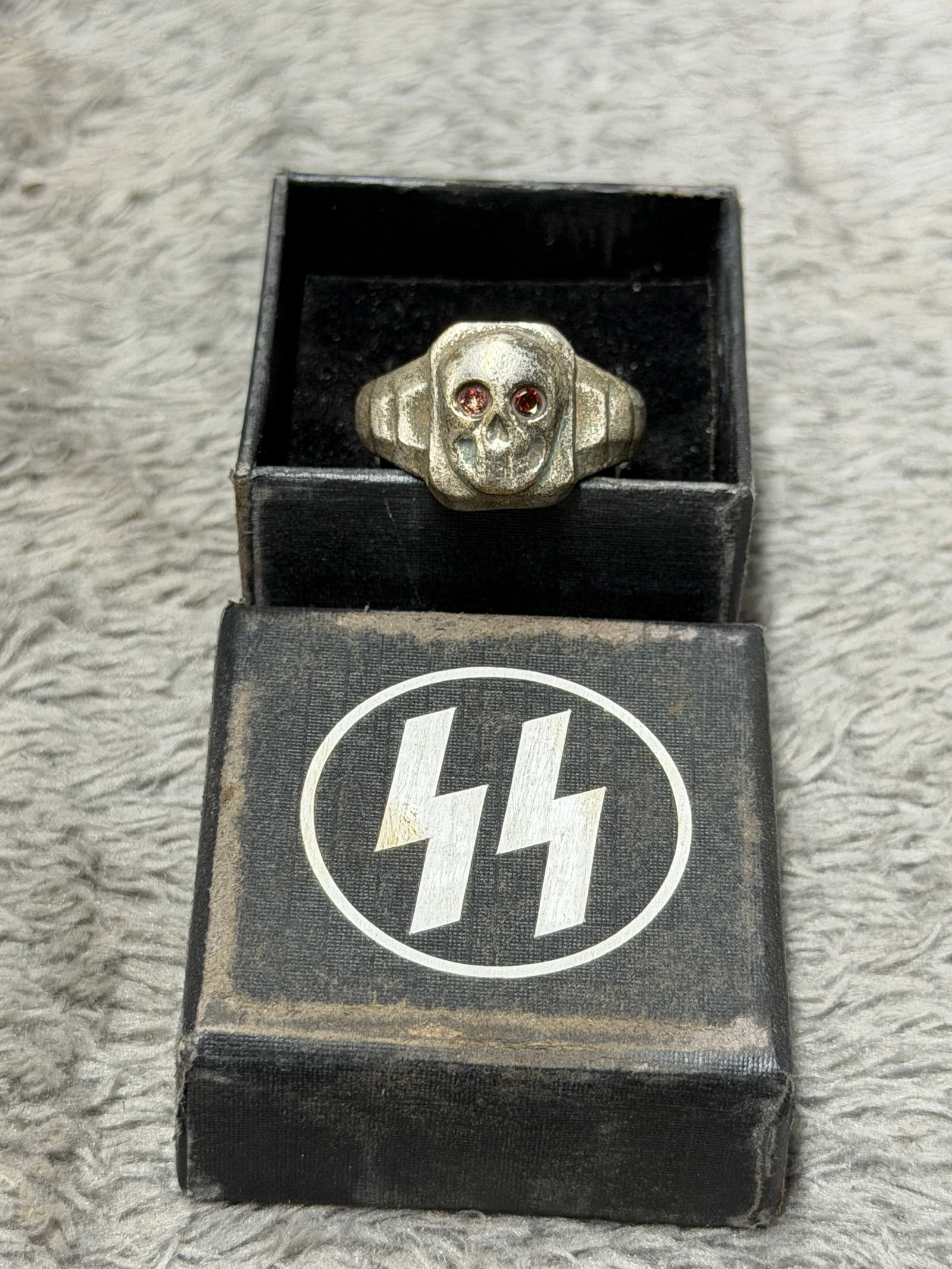 WWII German Waffen-SS Totenkopf Skull Ring with Red Stone Eyes & SS Presentation Box: Striking WWII German Waffen-SS themed finger ring featuring a prominent raised Totenkopf skull dominating the rectangular face, with deeply modeled eye sockets fitted with two red-colored stones that 
