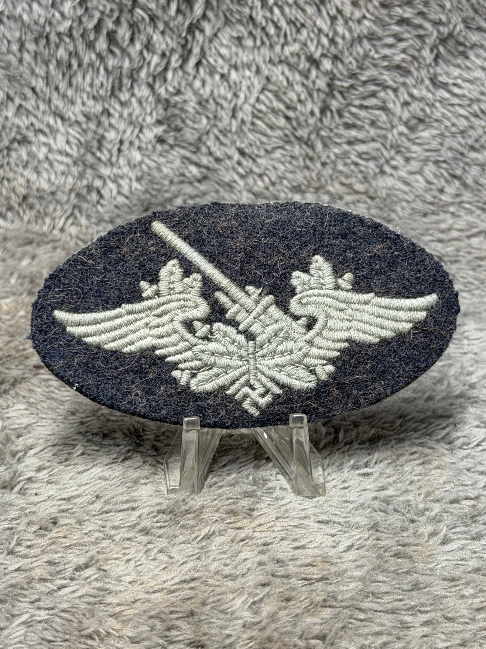WWII German Luftwaffe Anti-Aircraft Flak Artillery Gunner’s Sleeve Trade Badge: WWII German Luftwaffe embroidered sleeve trade badge featuring a detailed anti-aircraft artillery gun motif worked in silver-gray thread on an oval dark blue-gray wool backing. The central design depi