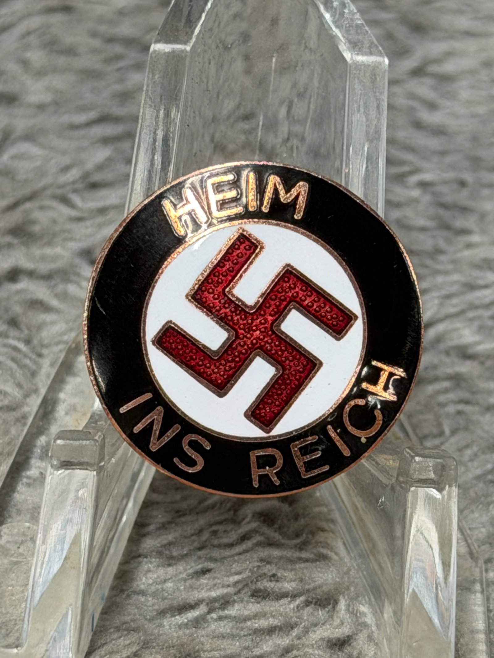 WWII German “Heim ins Reich?? NSDAP Enamel Membership Pin – R.R. Marked: WWII German political enamel pin displaying the inscription “HEIM INS REICH” surrounding a central red swastika on a white circular field, all set against a black enameled outer border with co
