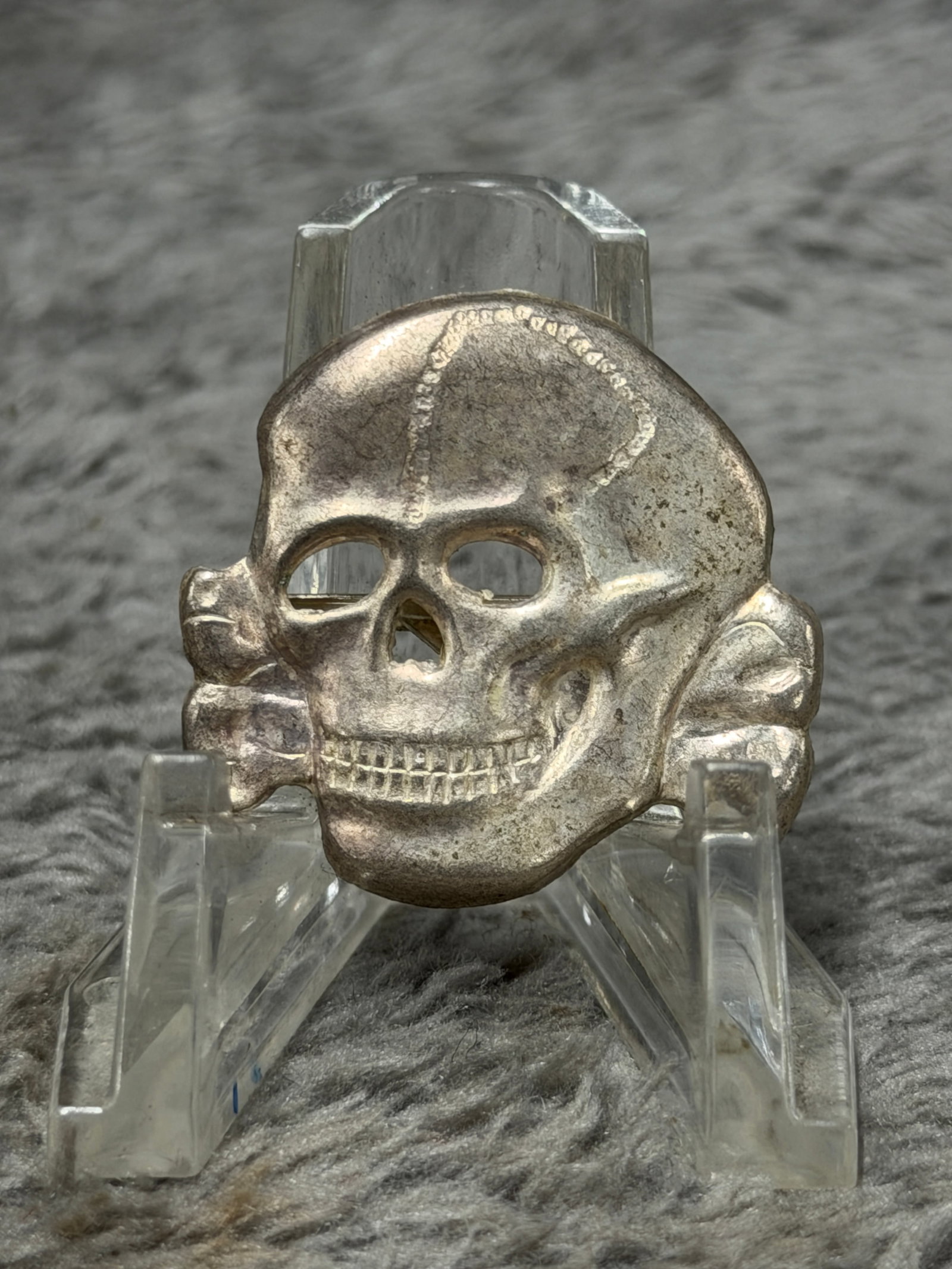 WWII German SS Totenkopf Death’s Head Skull Cap Insignia: WWII German SS Totenkopf skull insignia featuring the distinctive Death’s Head motif associated with SS headgear and insignia traditions. The piece is formed in a silver-colored metal with a promine