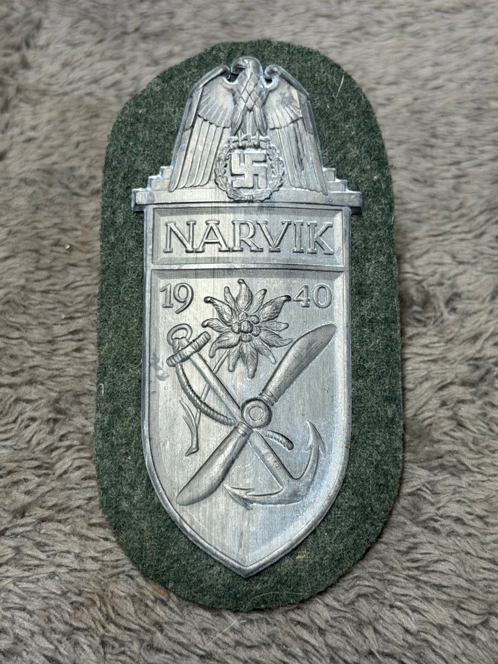 WWII German Narvik 1940 Campaign Shield with Kriegsmarine Naval Anchor & Edelweiss: WWII German Narvik 1940 campaign shield featuring a silver-tone metal insignia mounted to an oval field-gray/green cloth backing. The upper portion displays a spread-wing German eagle above a wreathed