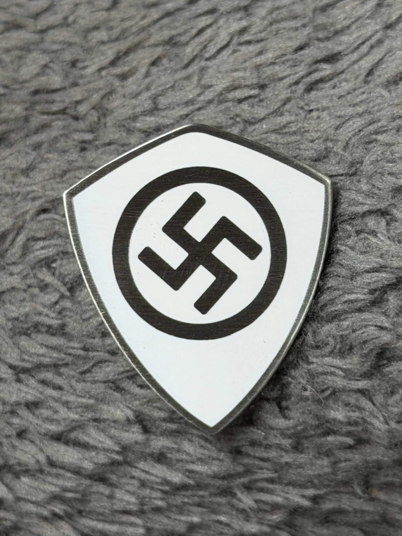 WWII German NSDAP Shield-Shaped Swastika Membership Pin Badge: WWII German political insignia fashioned in a distinctive shield-shaped form with a white enameled face dominated by a black swastika enclosed within a black circular border. A narrow exposed metal ri