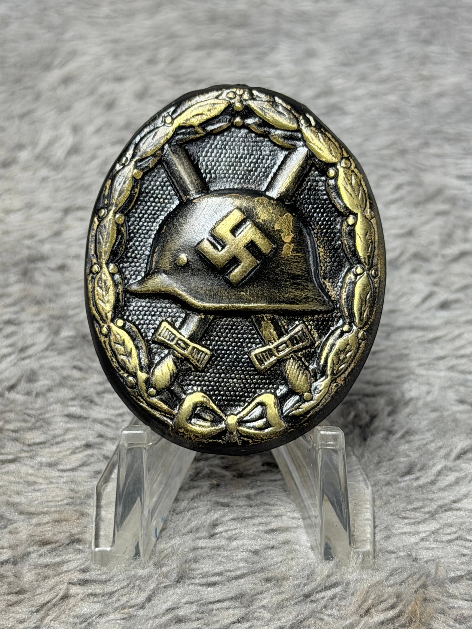 WWII German Black Wound Badge with Gold Highlighted Helmet & Swastika: WWII German oval Wound Badge featuring the characteristic central steel helmet bearing a raised swastika, set over crossed swords and surrounded by a detailed laurel wreath. The badge displays a predo