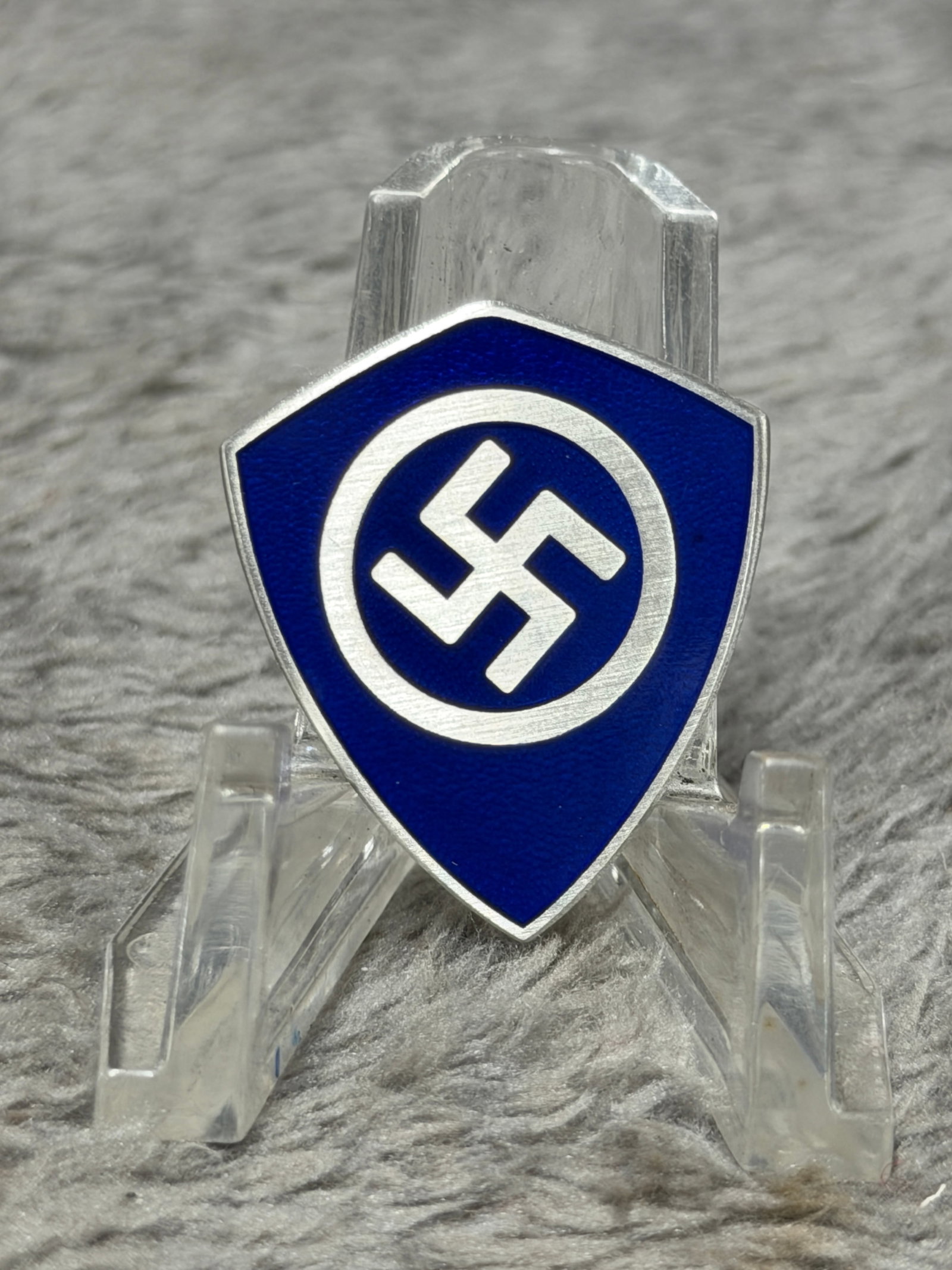 WWII German National Socialist Blue Enamel Swastika Shield Membership Badge – Ges. Gesch. Marked: Distinctive WWII German National Socialist membership or organizational badge featuring a shield-shaped metal body finished with deep blue enamel. The face displays a prominent silver-toned swastika e