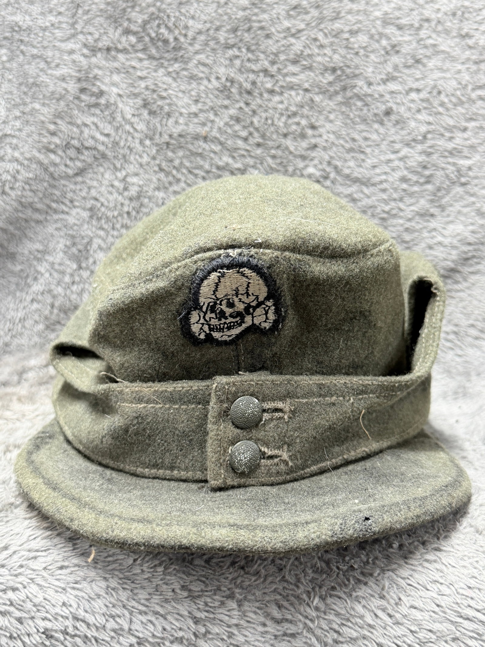 WWII German Waffen-SS M43 Field Cap with Totenkopf & Eagle Insignia: WWII German Waffen-SS M43-type field cap constructed of field-gray wool with a short cloth-covered visor, fold-down ear and neck panels, and a two-button front closure characteristic of this form of G