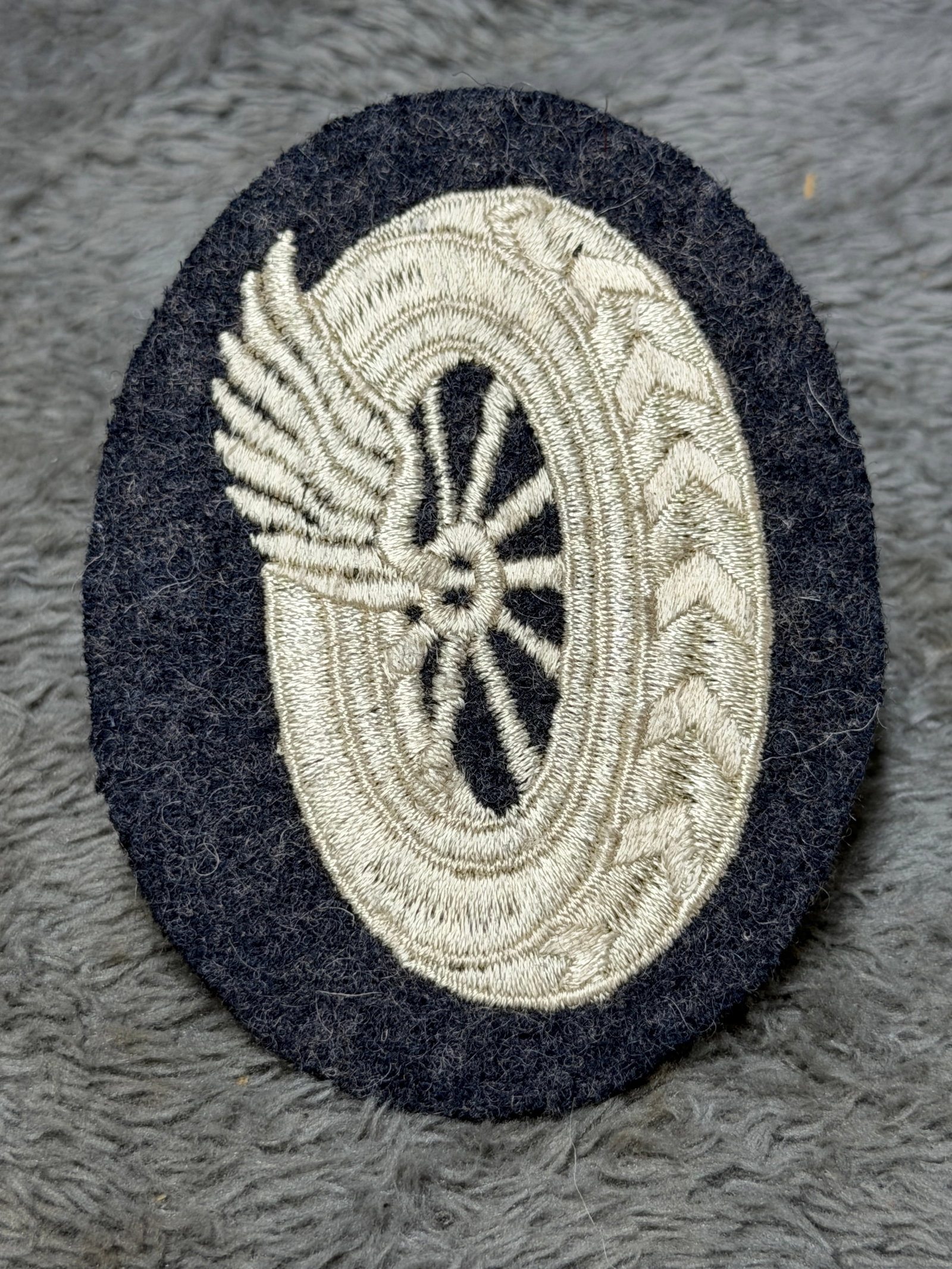 WWII German Luftwaffe Flight Personnel Embroidered Qualification Badge: WWII German Luftwaffe embroidered cloth qualification insignia featuring a finely rendered silver-gray winged propeller motif surrounded by an oval wreath, executed on a dark blue-black wool backing. 