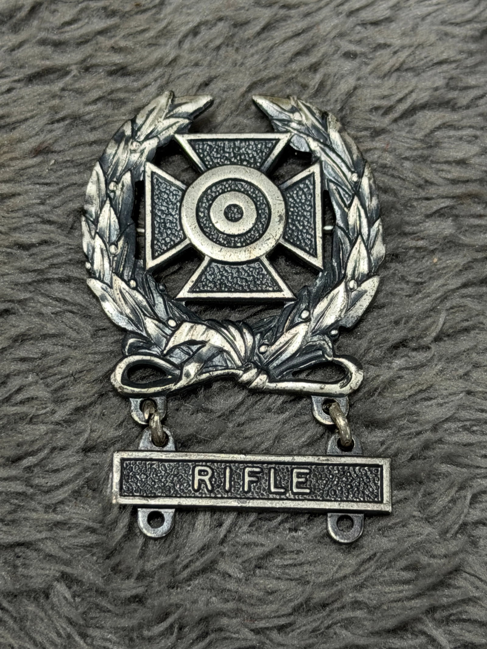 U.S. Military Sterling Rifle Marksmanship Qualification Badge – GI Marked: U.S. military marksmanship qualification badge featuring a prominent laurel wreath surrounding a Maltese-style cross with a concentric bullseye target at its center. The lower portion of the wreath is