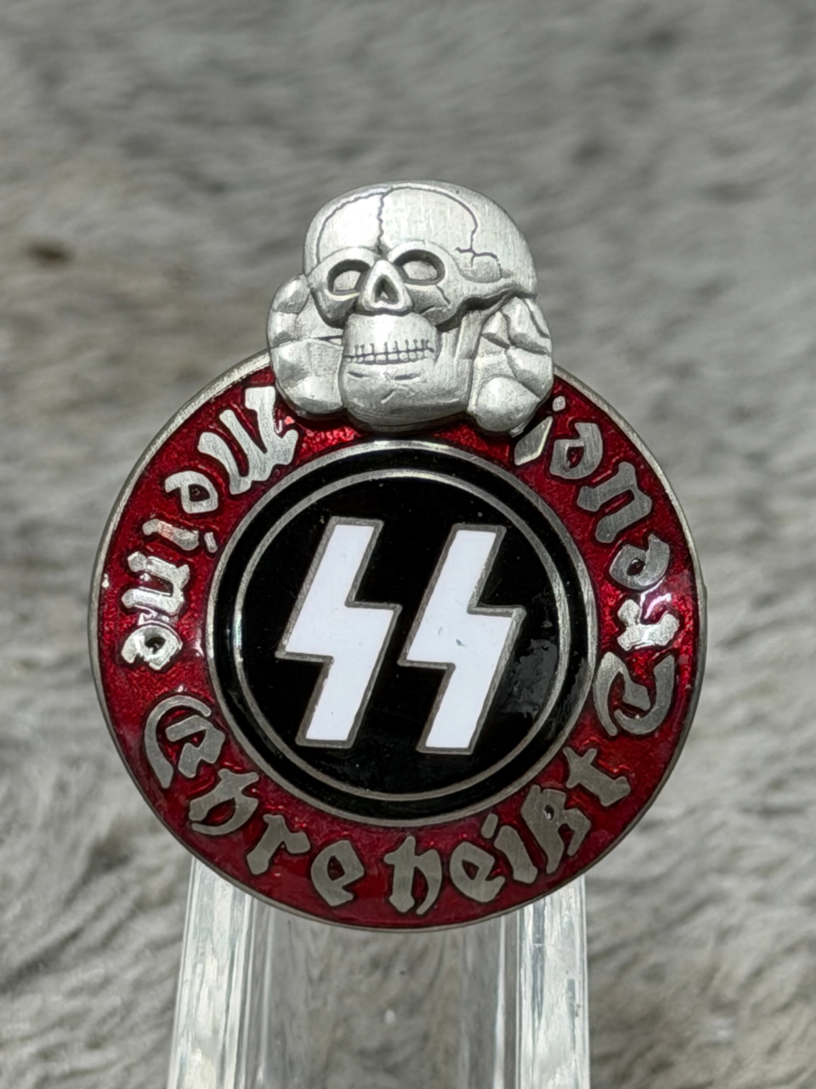 WWII German Waffen-SS “Meine Ehre Heißt Treue?? Totenkopf Membership Badge – RZM M1/42 Marked: WWII German Waffen-SS themed enamel membership badge featuring a striking circular red and black composition surmounted by a raised silver-colored Totenkopf skull emblem. The center displays the disti
