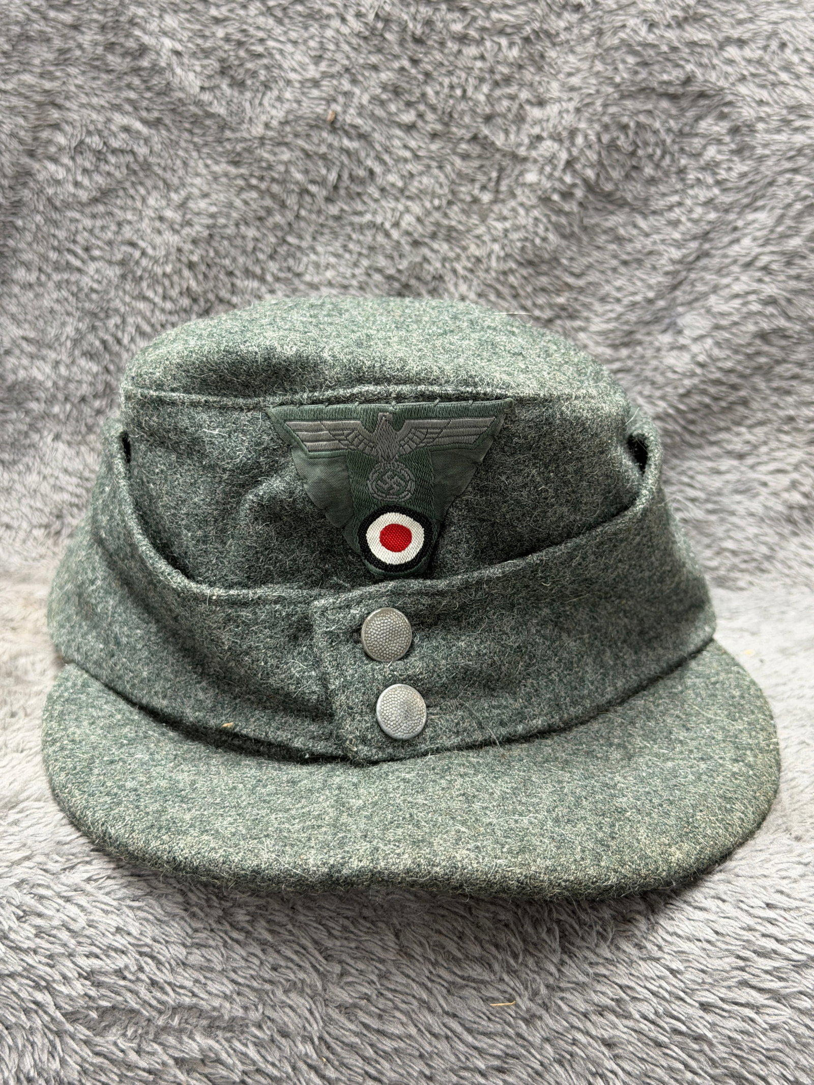WWII German Heer M43 Enlisted Field Cap with BeVo Eagle & Cockade: WWII German Heer M43 enlisted field cap constructed in field-gray wool with the characteristic short visor and fold-down side panels secured at the front by two pebbled metal buttons. The front displa