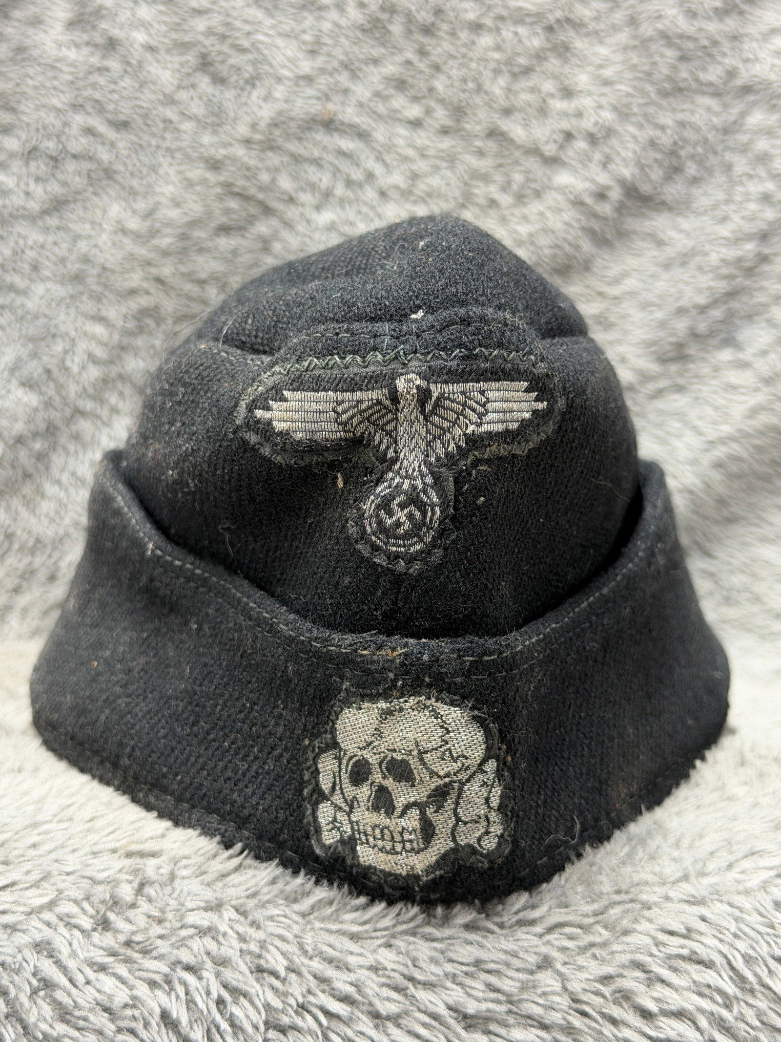 WWII German Waffen-SS M40 Black Overseas Field Cap with Totenkopf & Eagle Insignia: WWII German Waffen-SS black wool overseas field cap displaying prominent woven insignia to the front, including an SS-pattern eagle with outstretched wings positioned above a large Totenkopf death’s