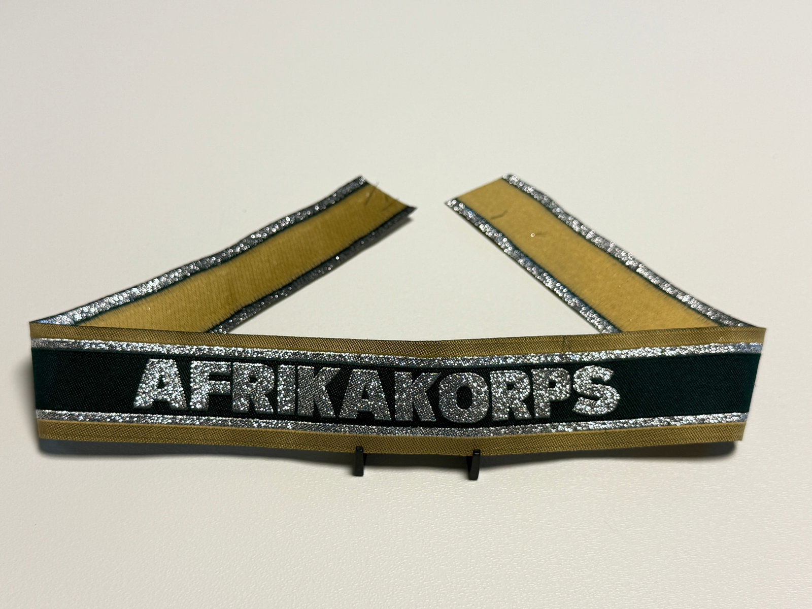 WWII German Afrika Korps “AFRIKAKORPS?? Officer’s Cuff Title: Striking WWII German Afrika Korps cuff title featuring a dark green central band prominently embroidered with the inscription “AFRIKAKORPS” in large silver-colored block lettering. The inscrip