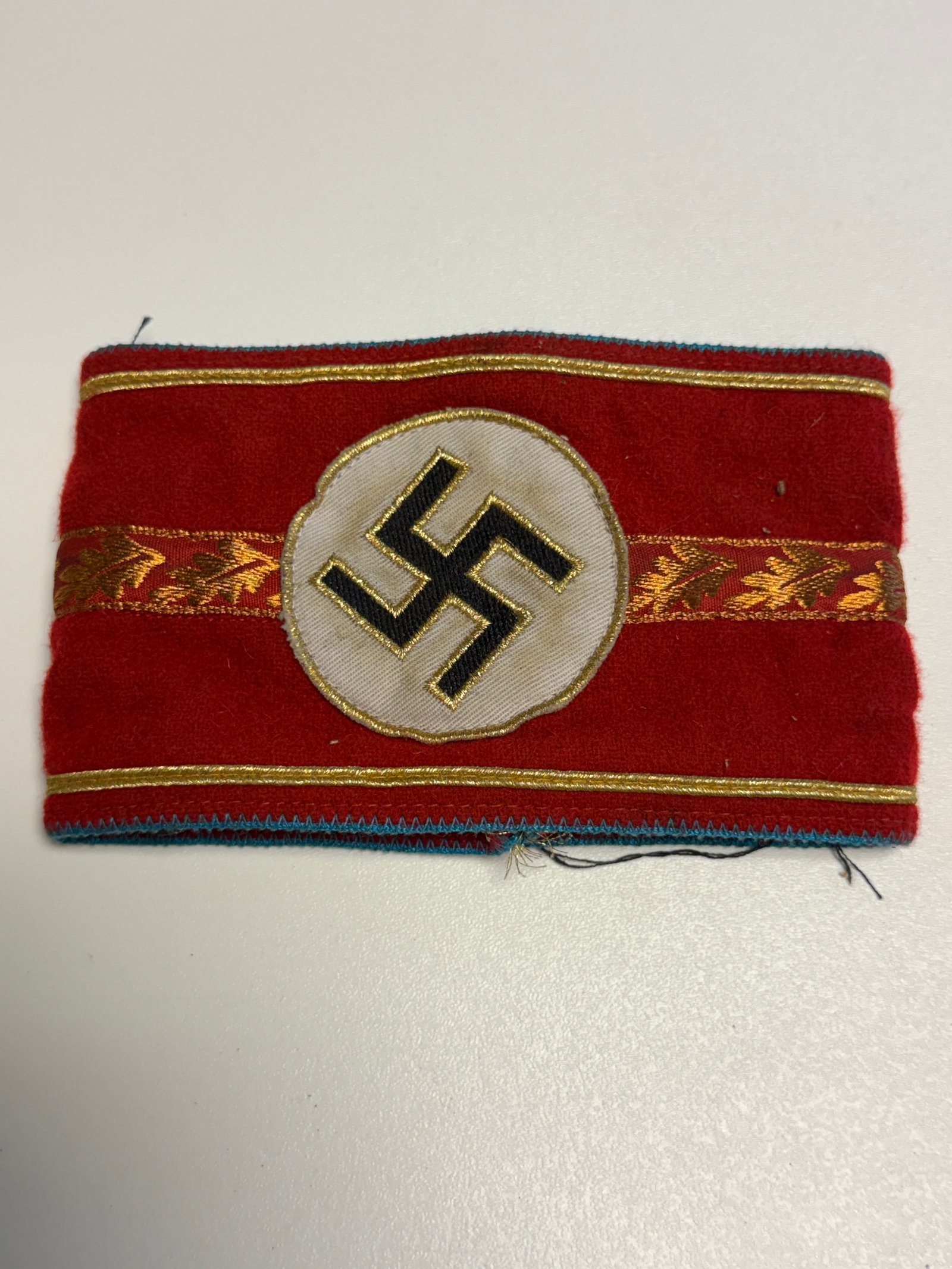 WWII German NSDAP Political Leader Gold-Trimmed Swastika Armband – RZM Label: WWII German NSDAP political organization armband constructed of red wool with an applied white circular field bearing a black embroidered swastika outlined in gold-colored thread. The armband is elabo