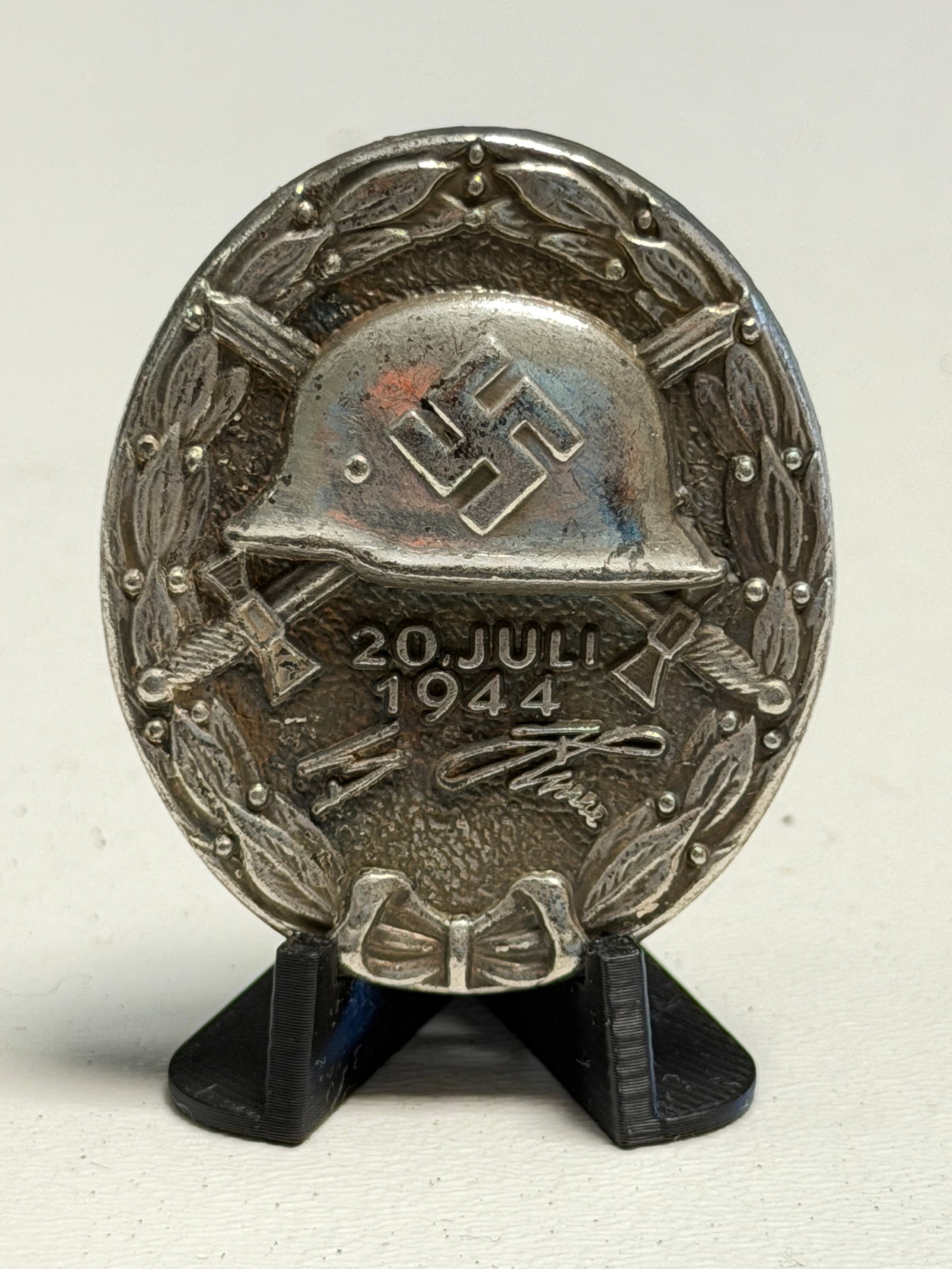 WWII German 20 July 1944 Wound Badge With Hitler Signature – “800 / 2?? Marked: WWII German commemorative Wound Badge dated “20 JULI 1944,” featuring a raised military helmet bearing a swastika above the prominent commemorative date, crossed swords, and a signature motif,