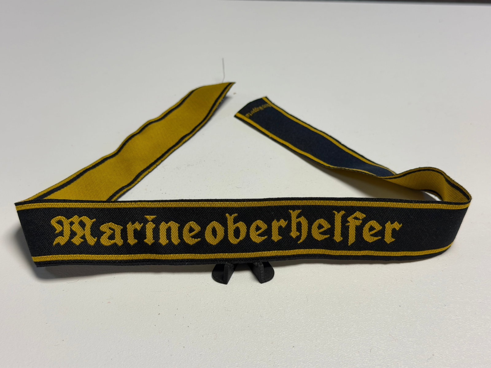 WWII German Kriegsmarine “Marineoberhelfer?? Naval Auxiliary Rank Tally: WWII German Kriegsmarine naval auxiliary textile insignia bearing the woven Gothic-script designation “Marineoberhelfer” in golden-yellow lettering against a dark navy-blue field, bordered abo