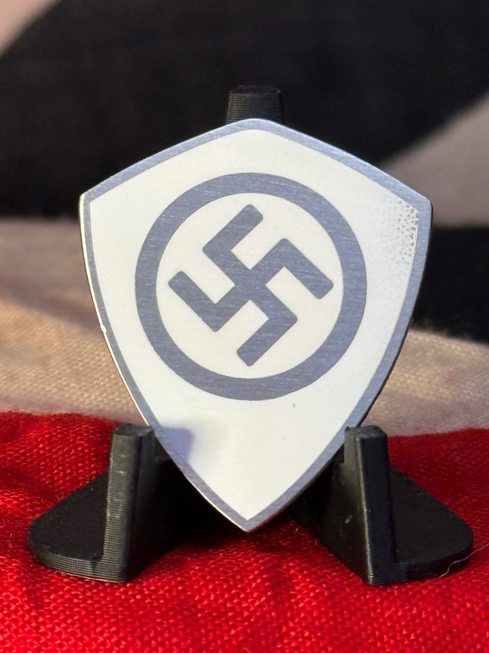 WWII German National Socialist Shield-Shaped Membership Badge – “Ges. Gesch.?? Marked: A shield-shaped German National Socialist pin badge featuring a prominent black swastika enclosed within a circular border against a light-colored field. The reverse is fitted with a vertically mounte