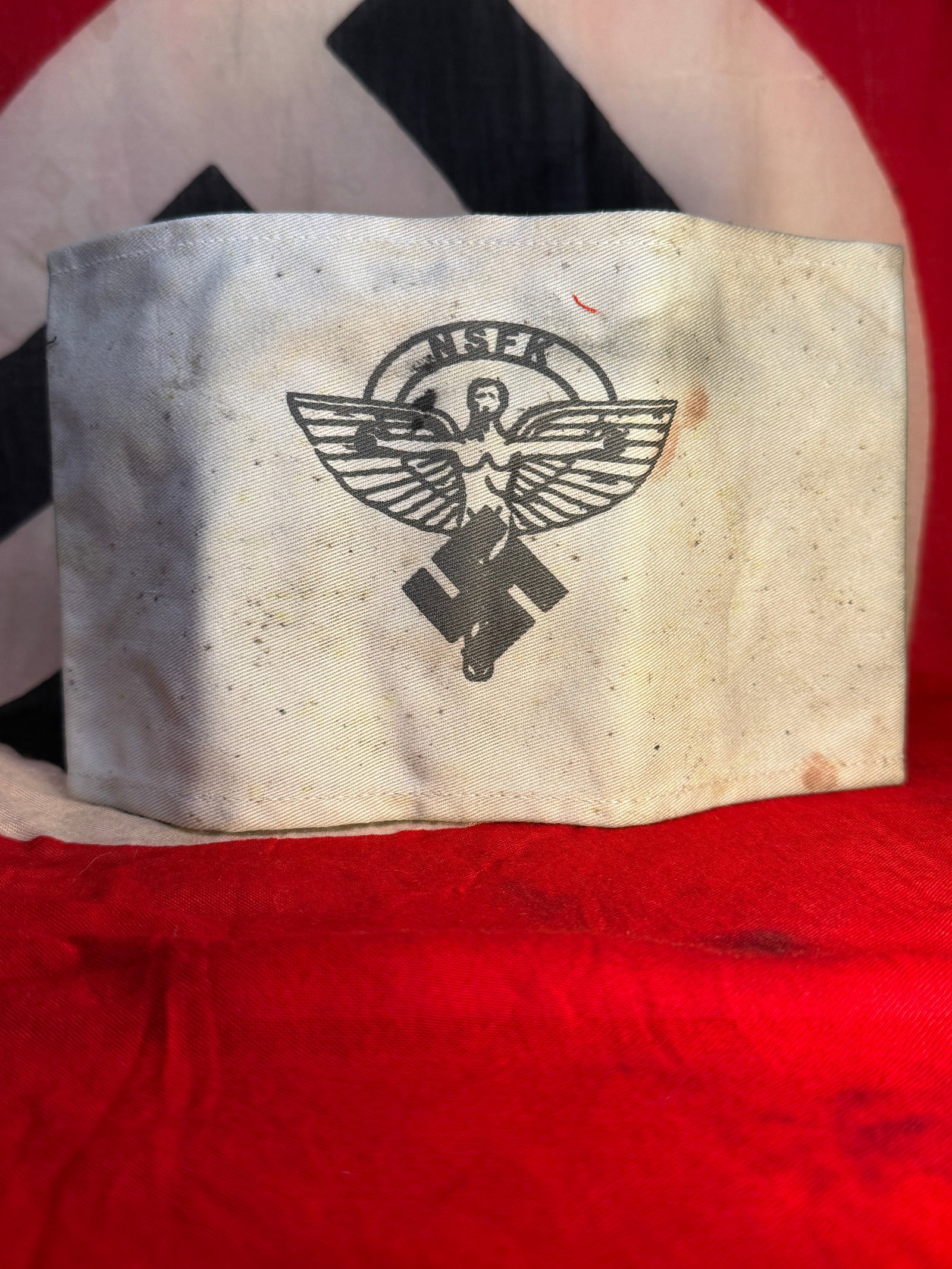 WWII German NSKK National Socialist Motor Corps Armband: A cloth armband associated with the NSKK (Nationalsozialistisches Kraftfahrkorps / National Socialist Motor Corps), constructed from light-colored fabric and displaying a printed NSKK emblem consistin