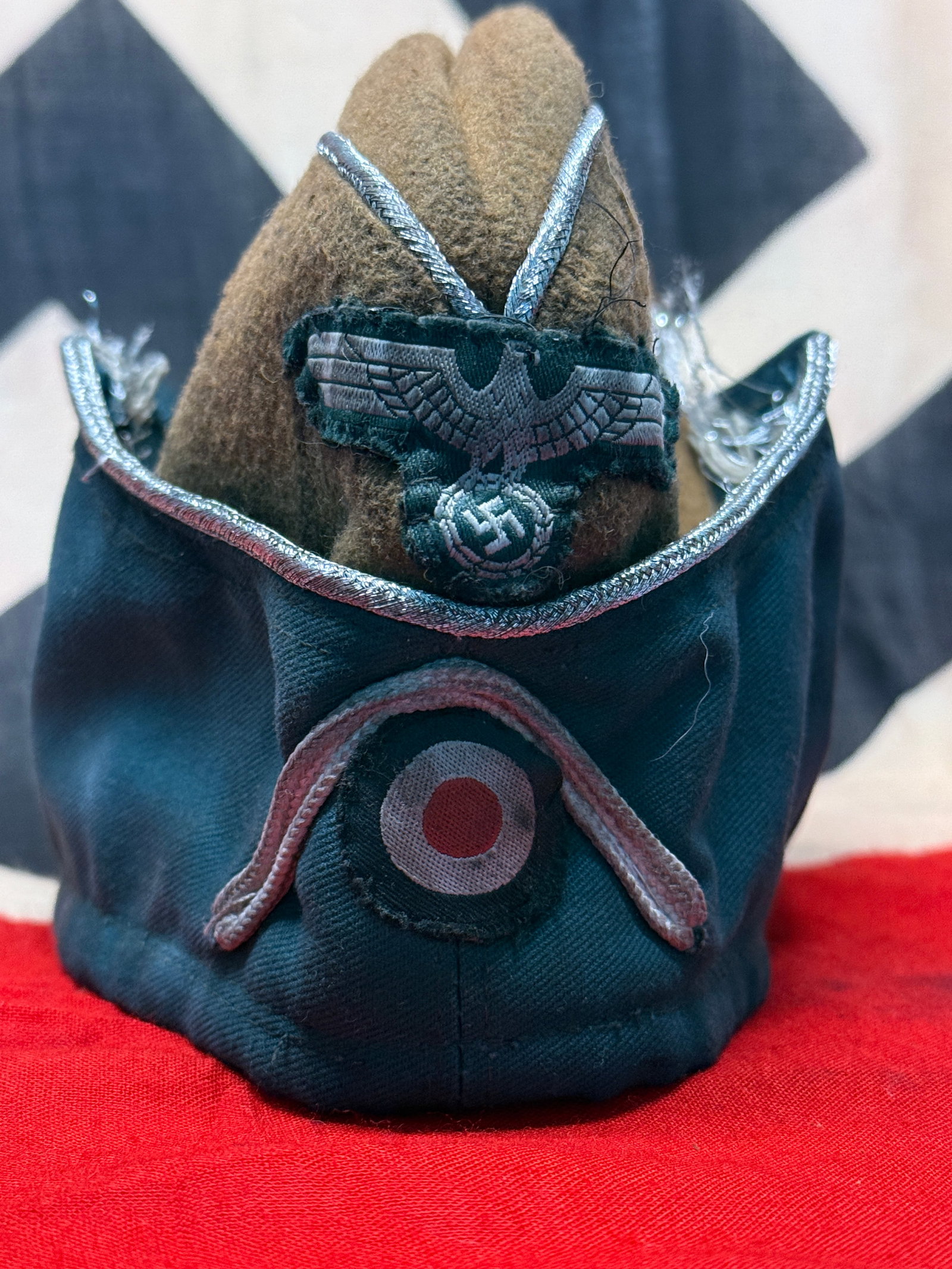 WWII German Heer Army Officer Overseas Side Cap with Silver Piping, Eagle & Cockade Insignia: WWII German Heer Army officer’s overseas side cap featuring a dark field-gray/blue-green cloth exterior with contrasting brown/tan upper section and silver-colored piping outlining the crown and upp