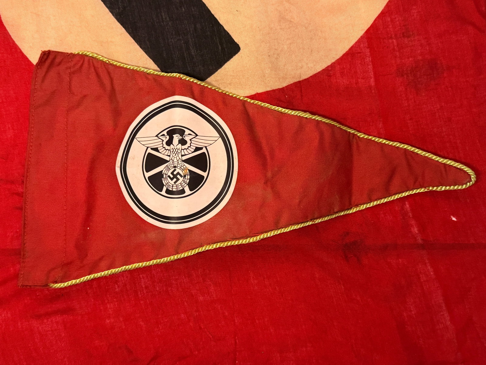 WWII German NSKK National Socialist Motor Corps Vehicle Pennant: A triangular red vehicle pennant associated with the WWII German NSKK, or Nationalsozialistisches Kraftfahrkorps (National Socialist Motor Corps). The pennant features a large circular emblem displayi
